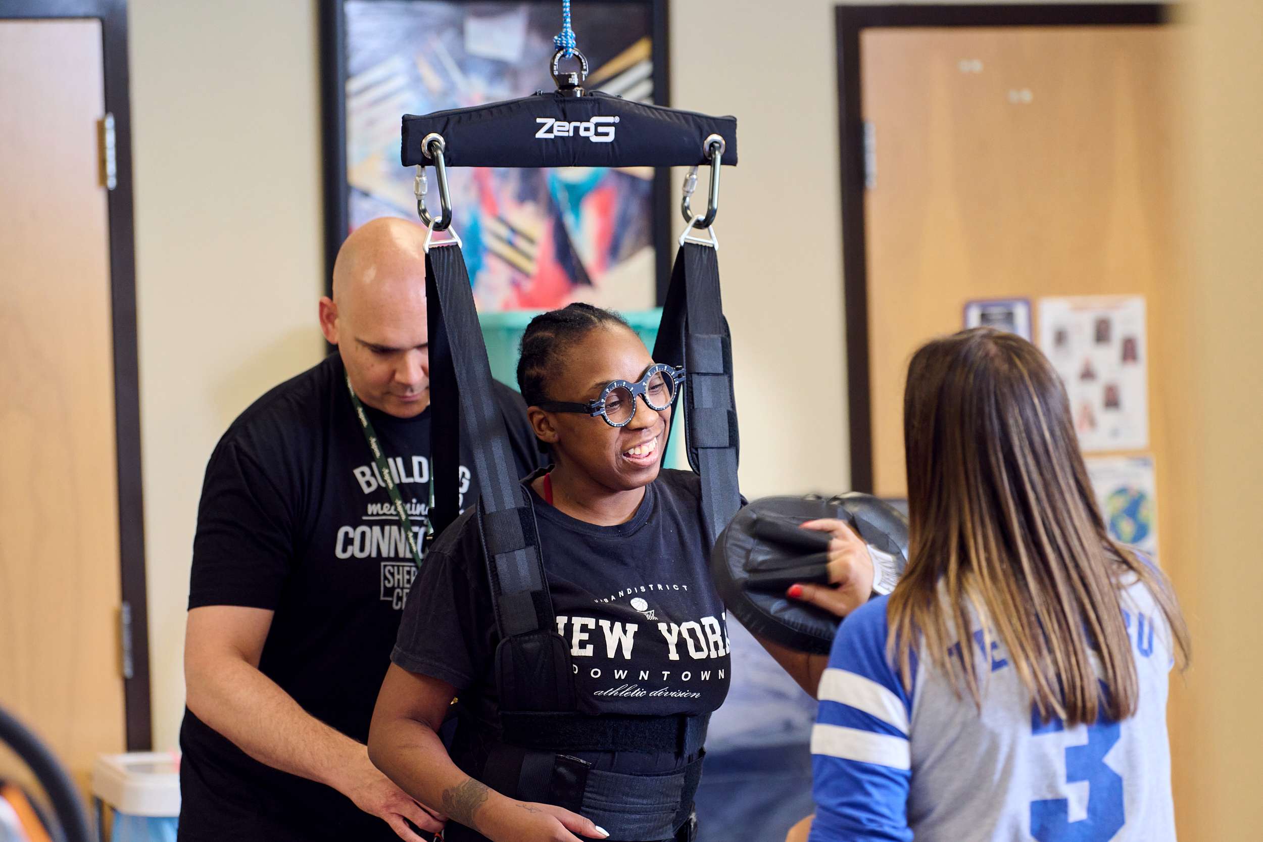 A man assists a woman using a harness for support in a therapy session. Another person stands nearby, wearing a blue and white jersey. They are in a room with closed doors and artwork on the walls.