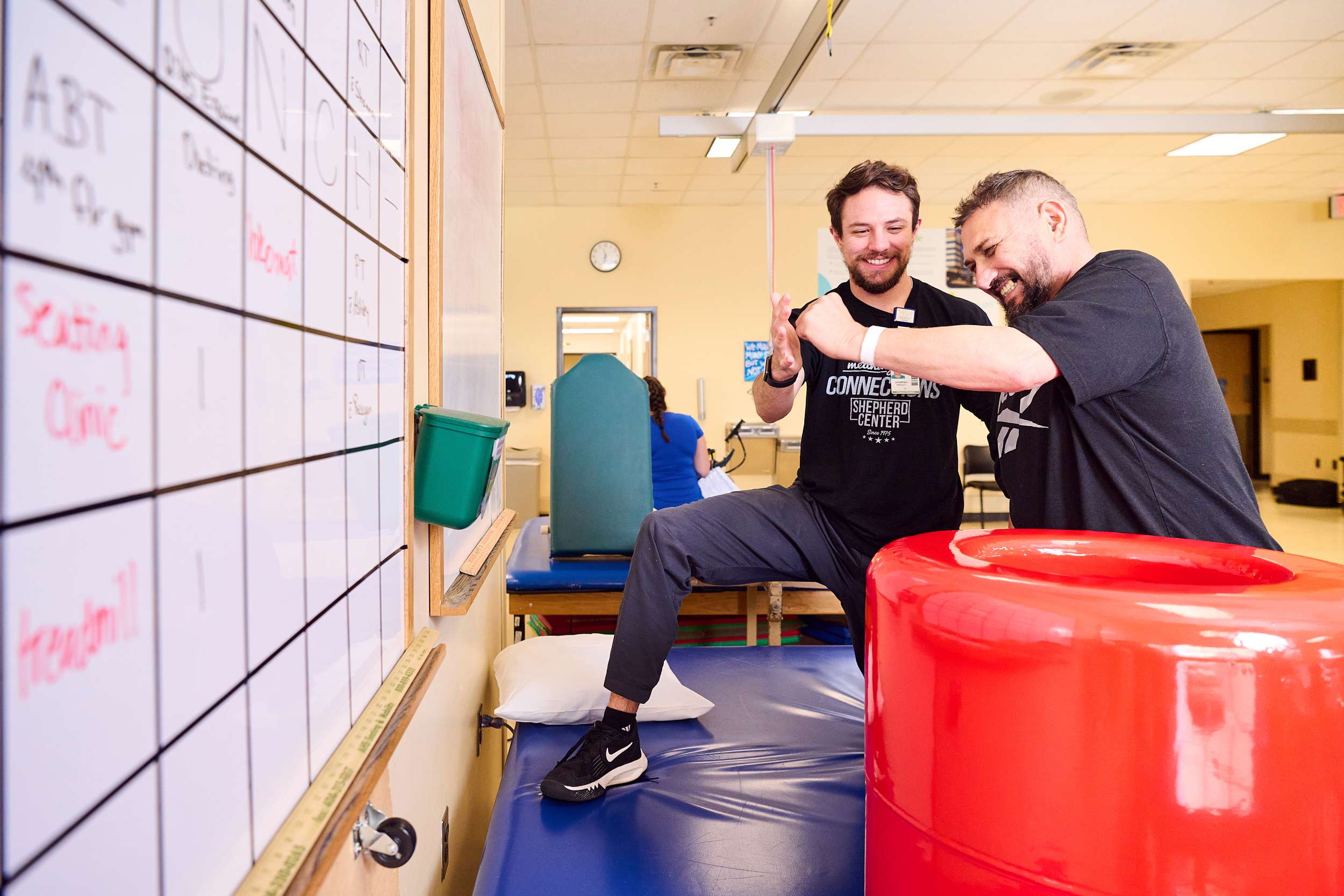 Male patient punches towards physical therapists' outstretched hand while kneeling on a blue table in the therapy gym.