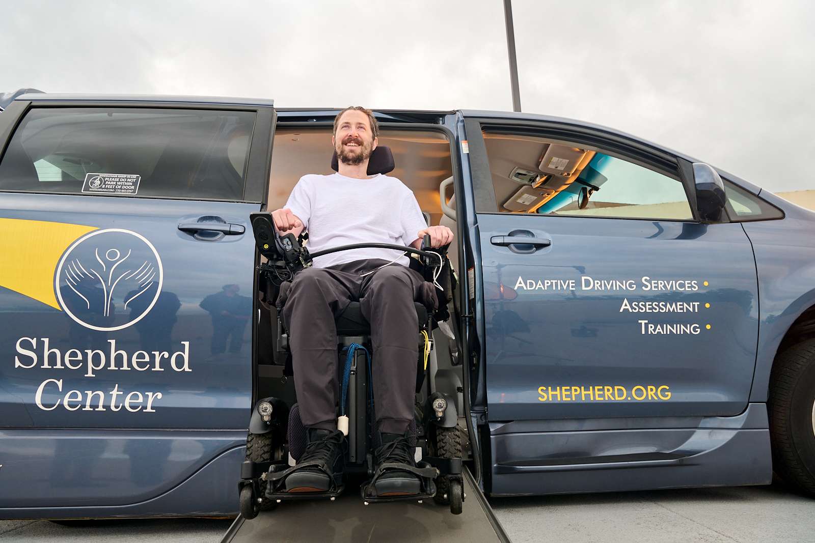 A person in a wheelchair smiles while exiting an accessible van via a ramp. The van is labeled with the Shepherd Center logo and offers services like adaptive driving, assessment, and training.