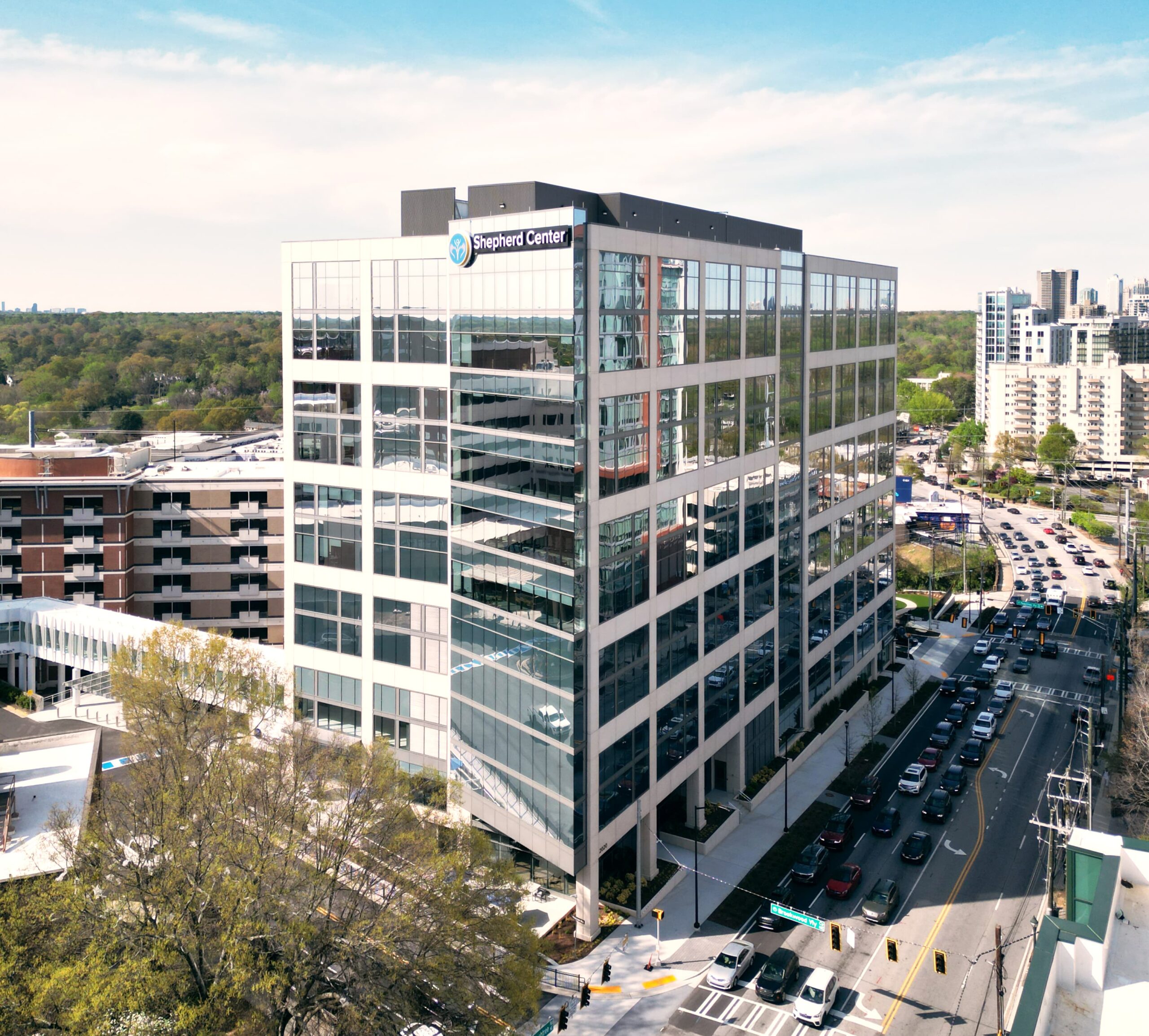 A modern, multi-story office building with large glass windows stands at a street corner. People walk on the sidewalks, and cars are on the road. The sky is clear and blue, and nearby trees add greenery to the urban scene.