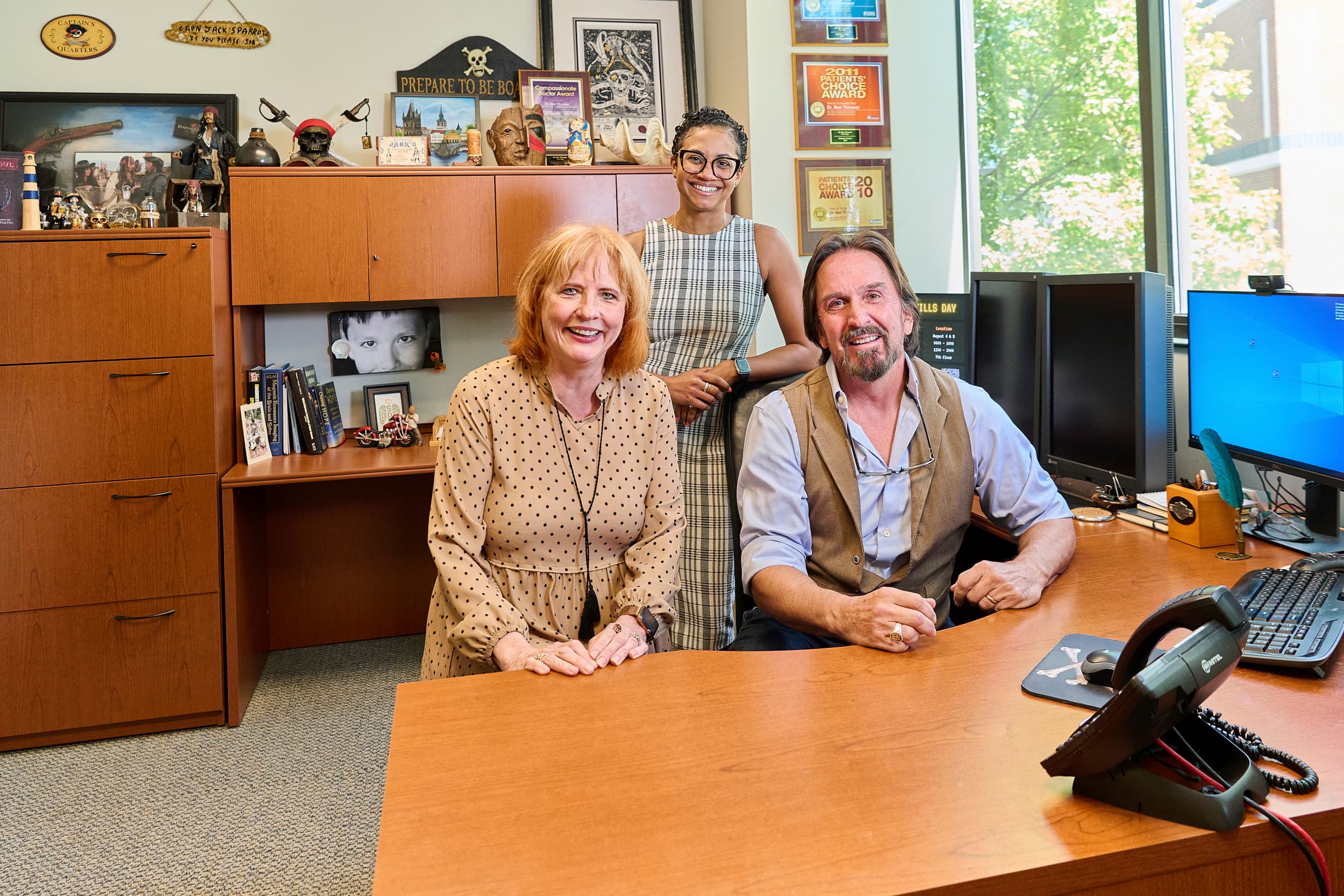 Three people are in an office setting. A man is seated at a desk with a computer, while two women stand nearby. The office is decorated with shelves, books, and photos. There is a window showing greenery outside. All are smiling.