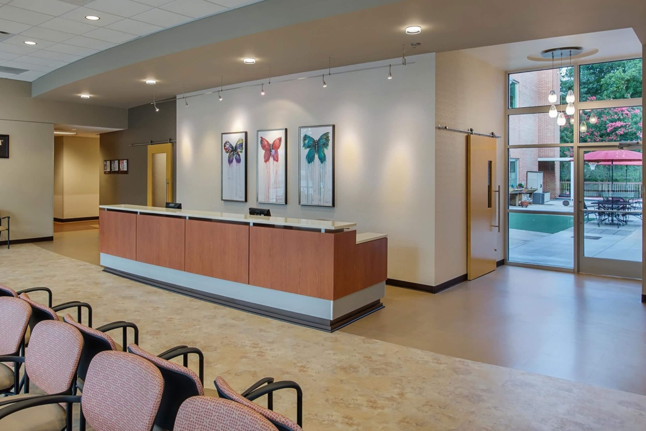 Reception area with a curved wooden desk and several chairs. Three butterfly-themed artworks hang on the cream-colored wall behind the desk. Large windows on the right let in natural light, revealing an outdoor area with tables and umbrellas.