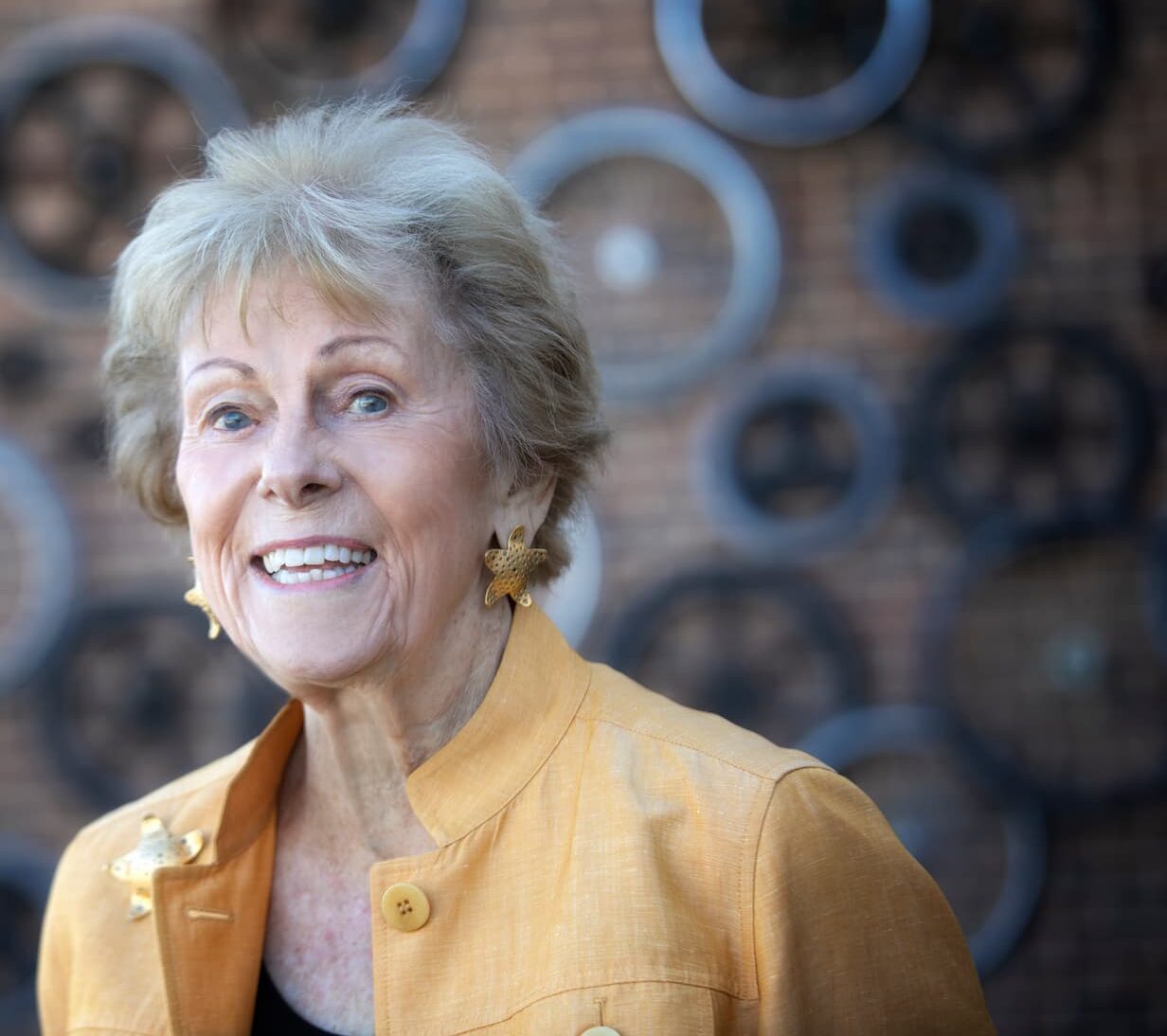 A woman with light hair smiles warmly, wearing a yellow jacket and star-shaped earrings. She stands in front of a brick wall decorated with circular metal designs of various sizes.