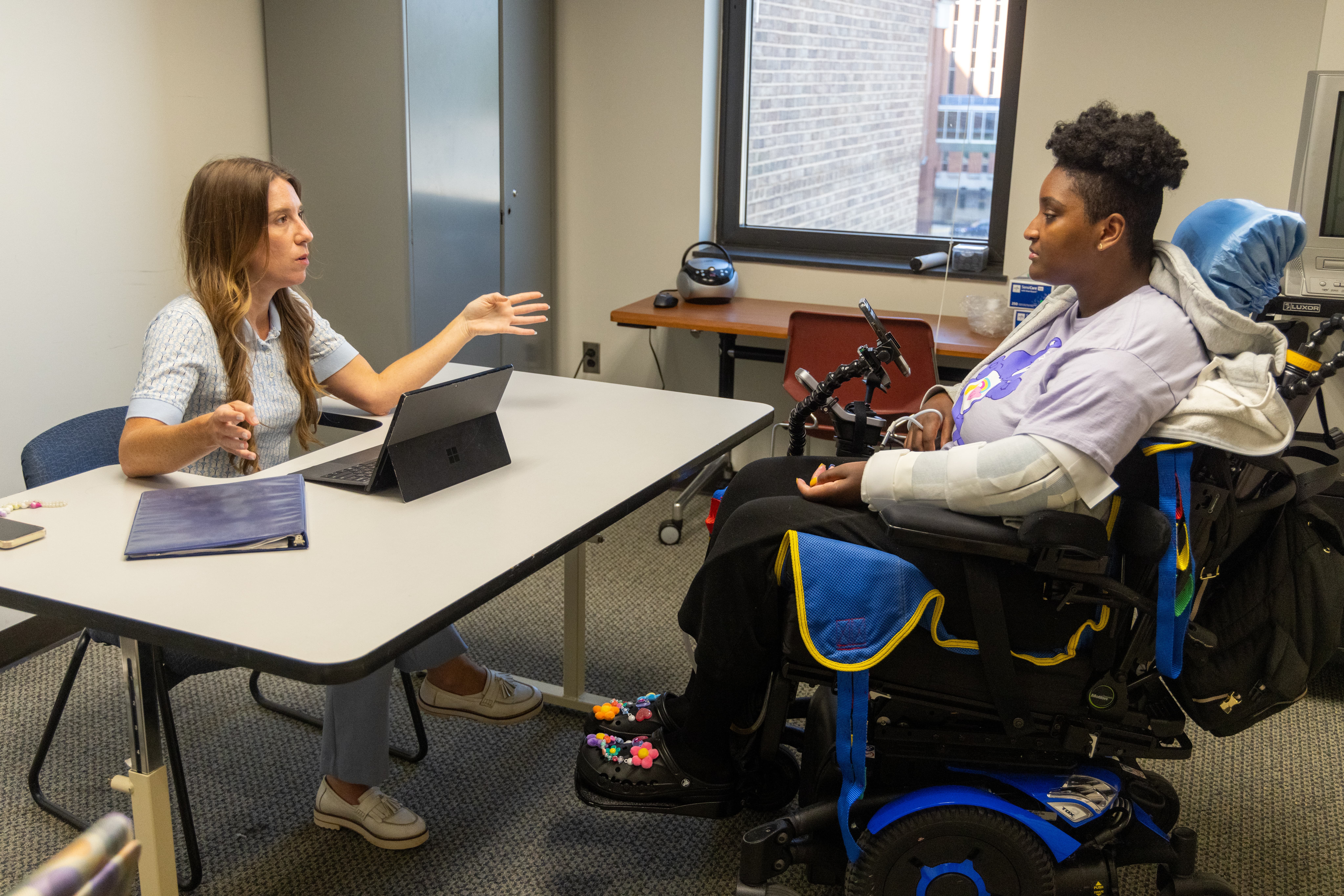 A woman speaks with another woman in a power wheelchair across a table in an office. The woman in the wheelchair has her arm in a cast and medical equipment attached to her chair. Both appear to be engaged in conversation.