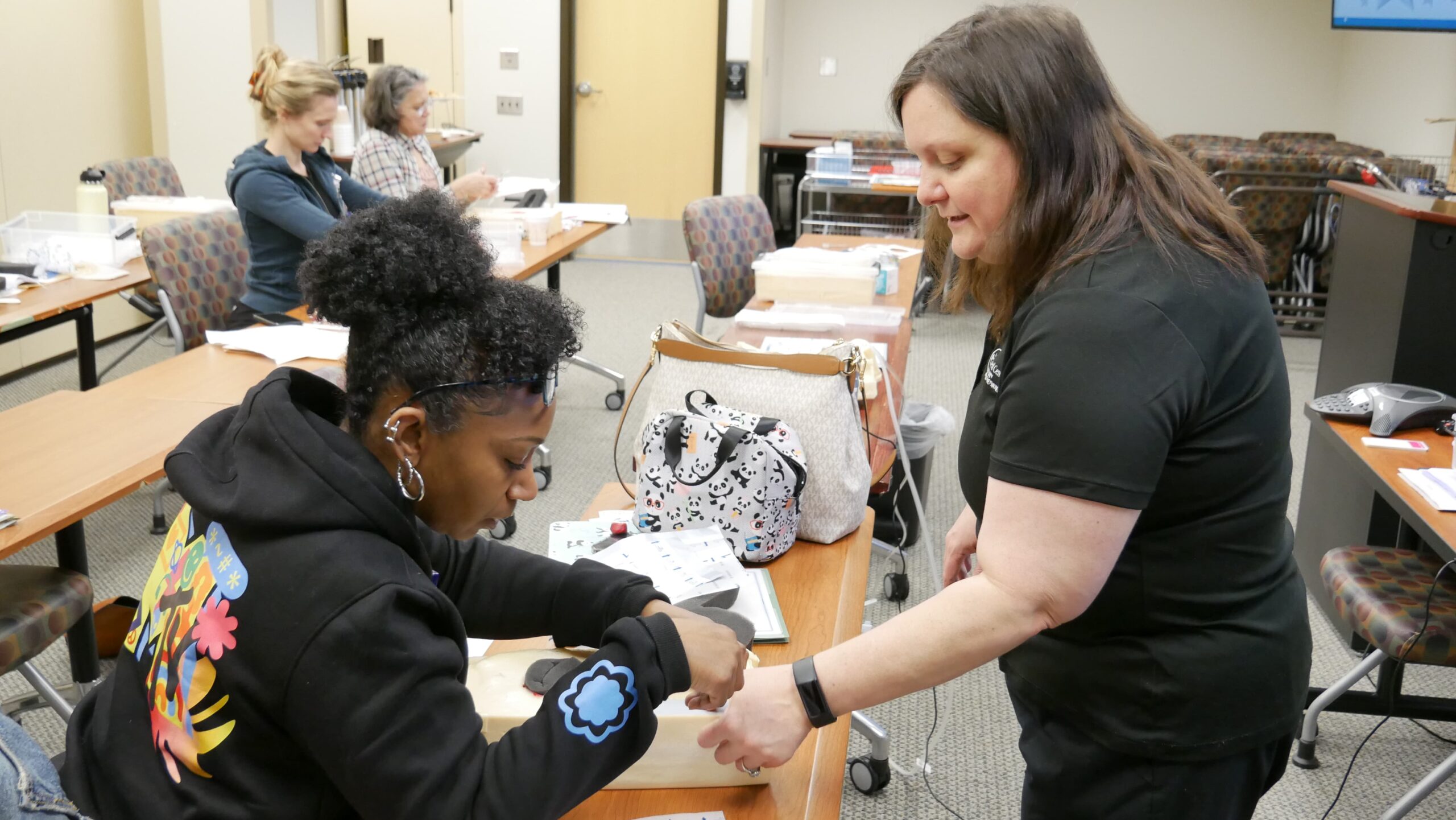 A woman assists another woman who is seated. They are in a classroom with several tables and chairs. Other people are visible in the background, engaging in various activities. Bags and materials are on the tables.