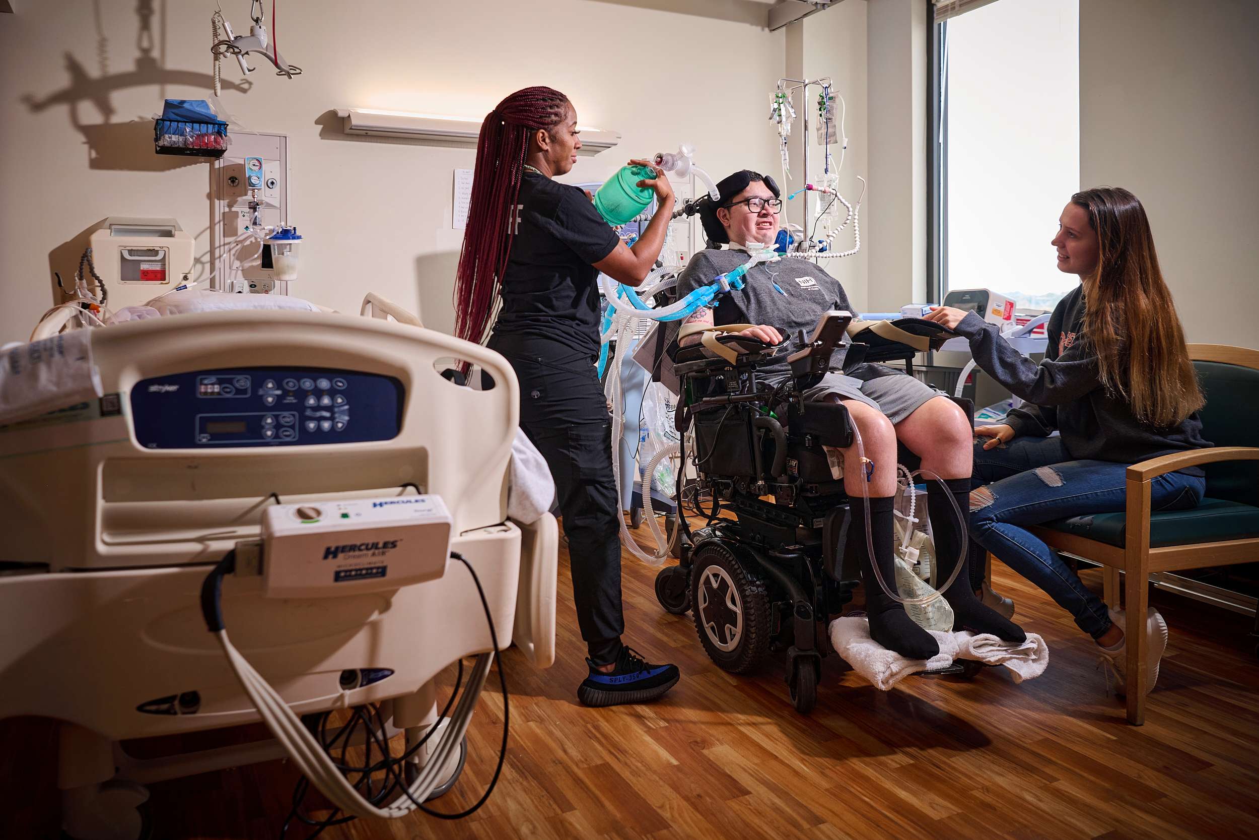A respiratory therapist attends to a patient in a motorized wheelchair in a hospital room. The patient's wife is seated nearby, engaging with the patient.
