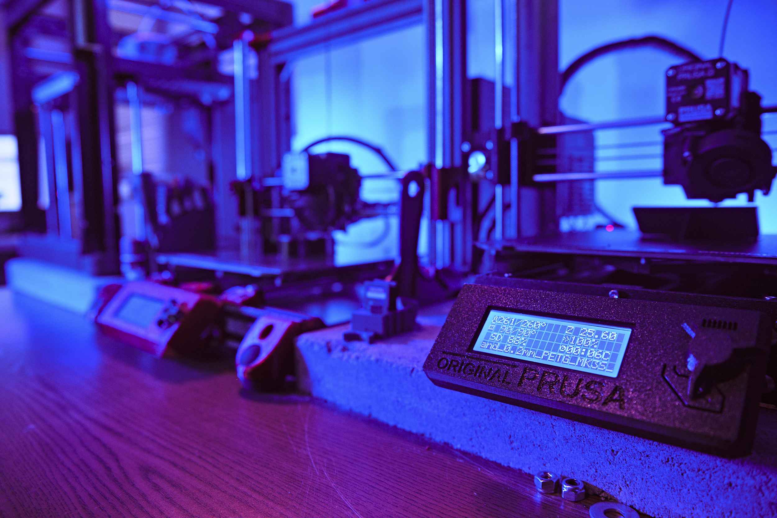 Close-up of a 3D printer on a wooden table with a blue-lit background. The printer's LCD screen displays settings and information. Multiple 3D printers are visible in the background, creating a high-tech workshop atmosphere.