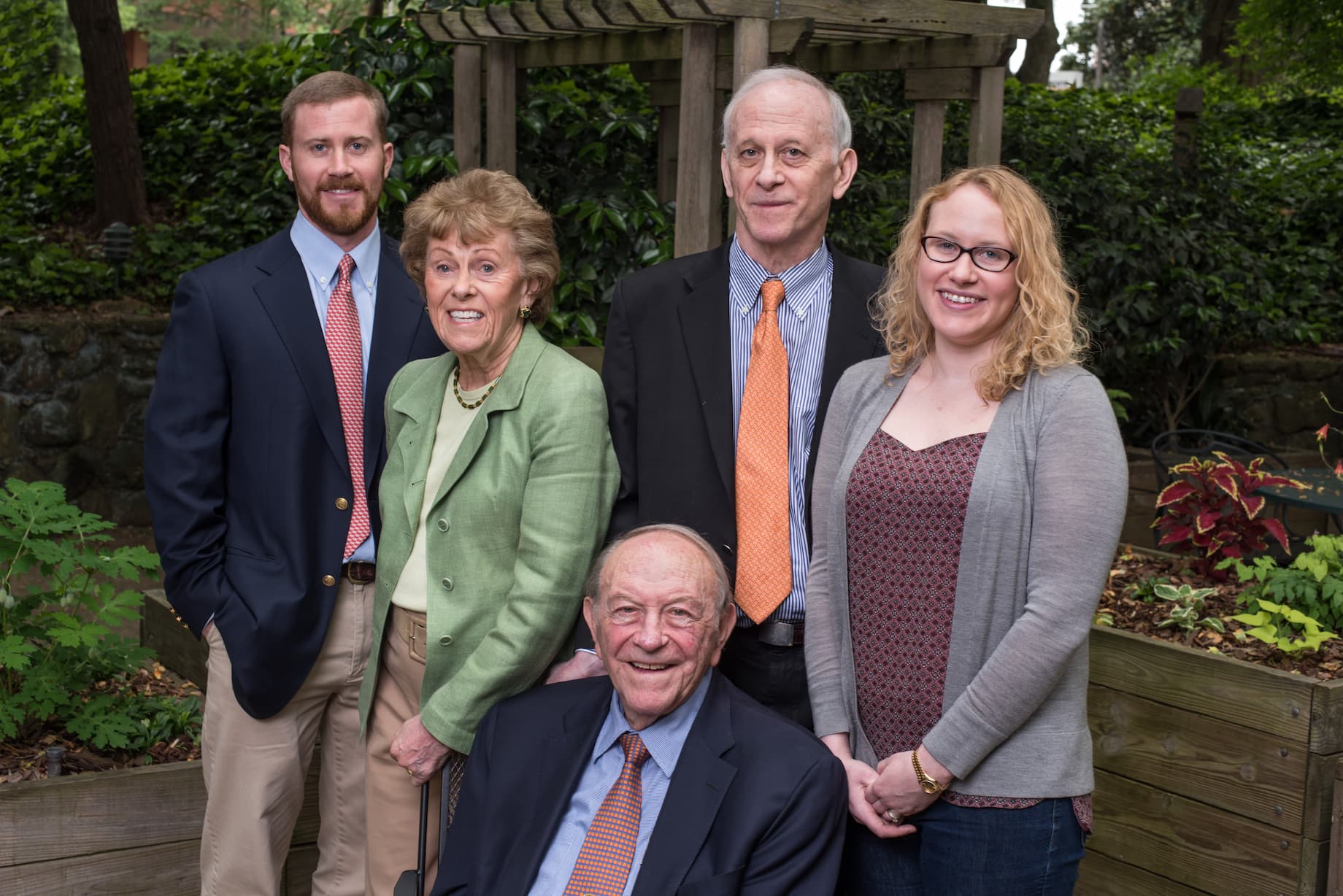 A group of five people, four standing and one seated, are outdoors in a garden setting. They are dressed in semi-formal attire. There is greenery and a wooden structure in the background.