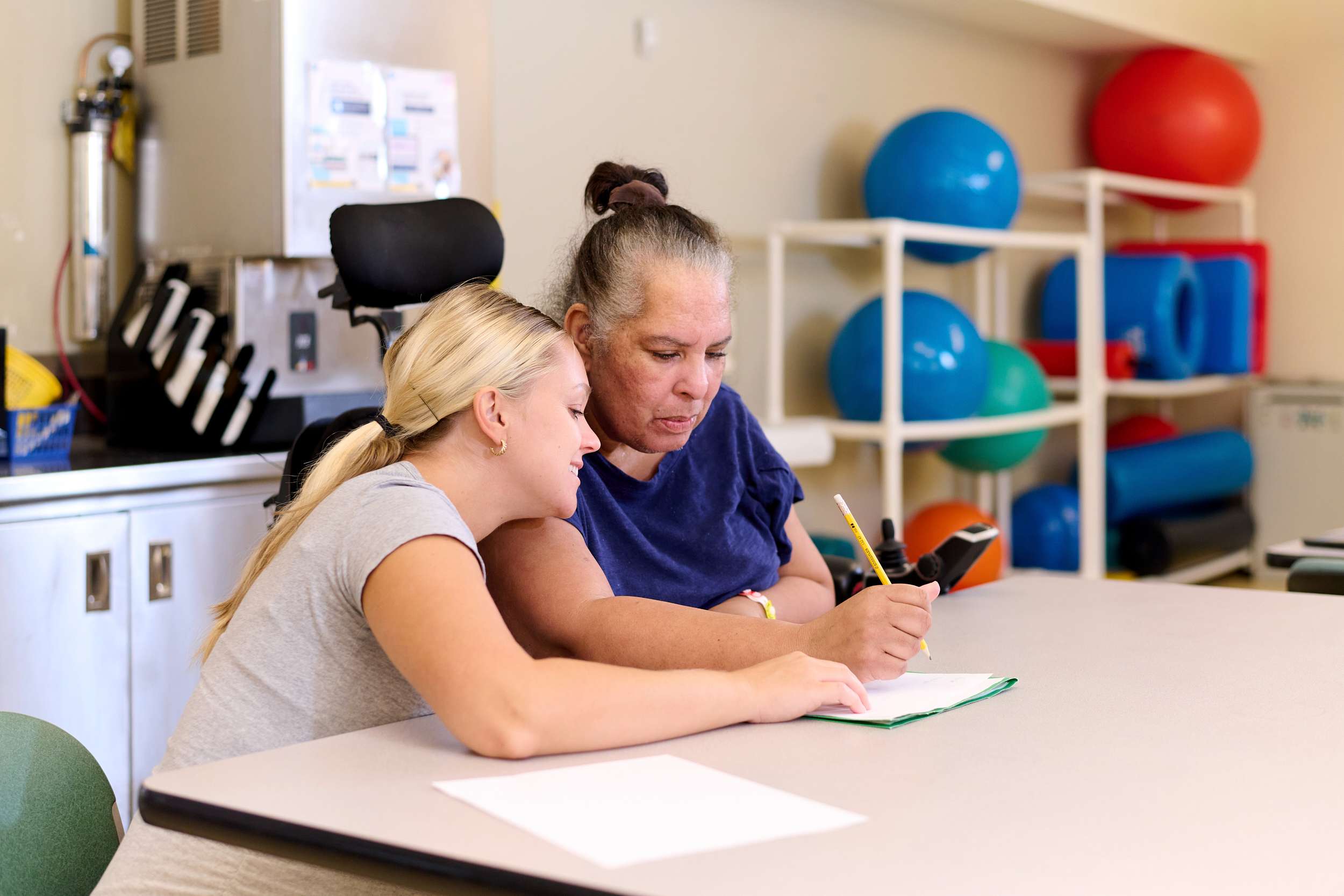 A woman with gray hair and a blue shirt is sitting at a table, writing on paper with a pencil. A blonde woman in a gray shirt leans over, smiling as they work together. Colorful exercise balls are in the background.