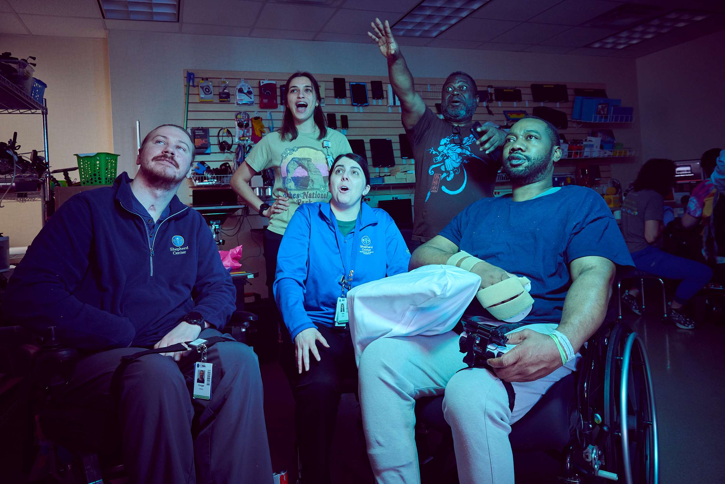 A group of five people, some using wheelchairs, are in a room with shelves in the background. They are looking towards the ceiling, with expressions of excitement and wonder. One person raises an arm enthusiastically. The lighting casts a blue tone.