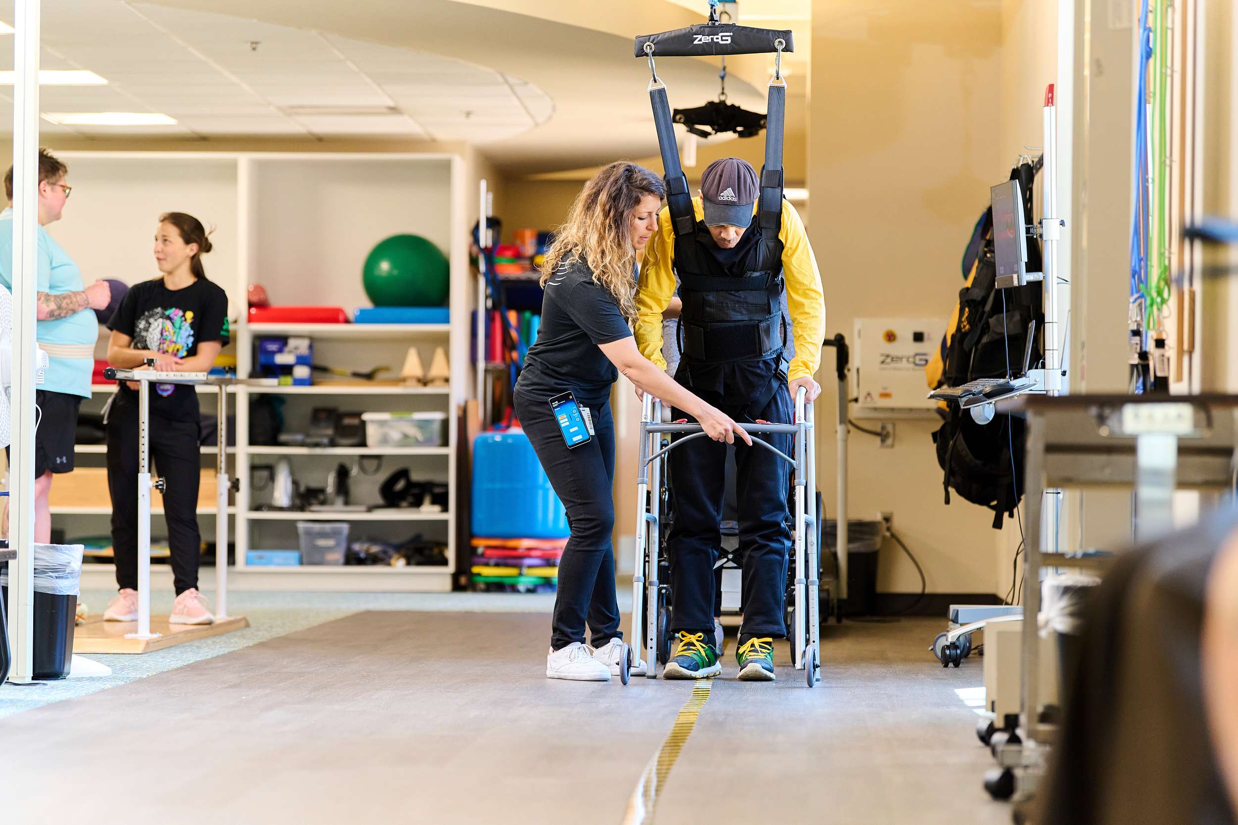 A therapist assists a man using a harness and walking frame in a rehabilitation center. The man wears a cap and casual clothes. In the background, two people stand near a shelf with exercise equipment and colorful balls.