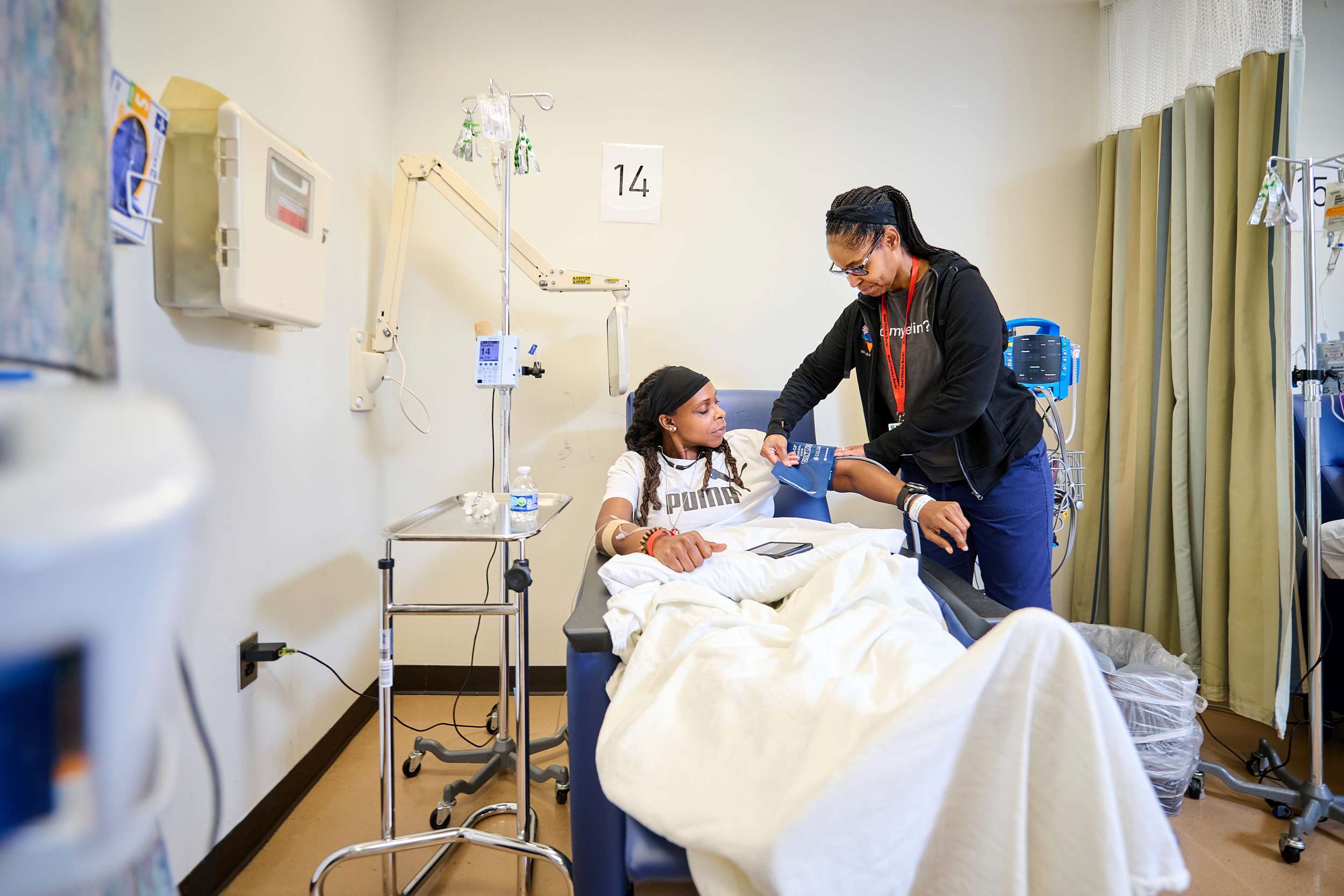 A patient lies in a hospital bed receiving care from a healthcare worker. The worker, standing beside the bed, is adjusting a device on the patient's arm while a medical monitor and IV stand are nearby. Both appear focused and engaged.