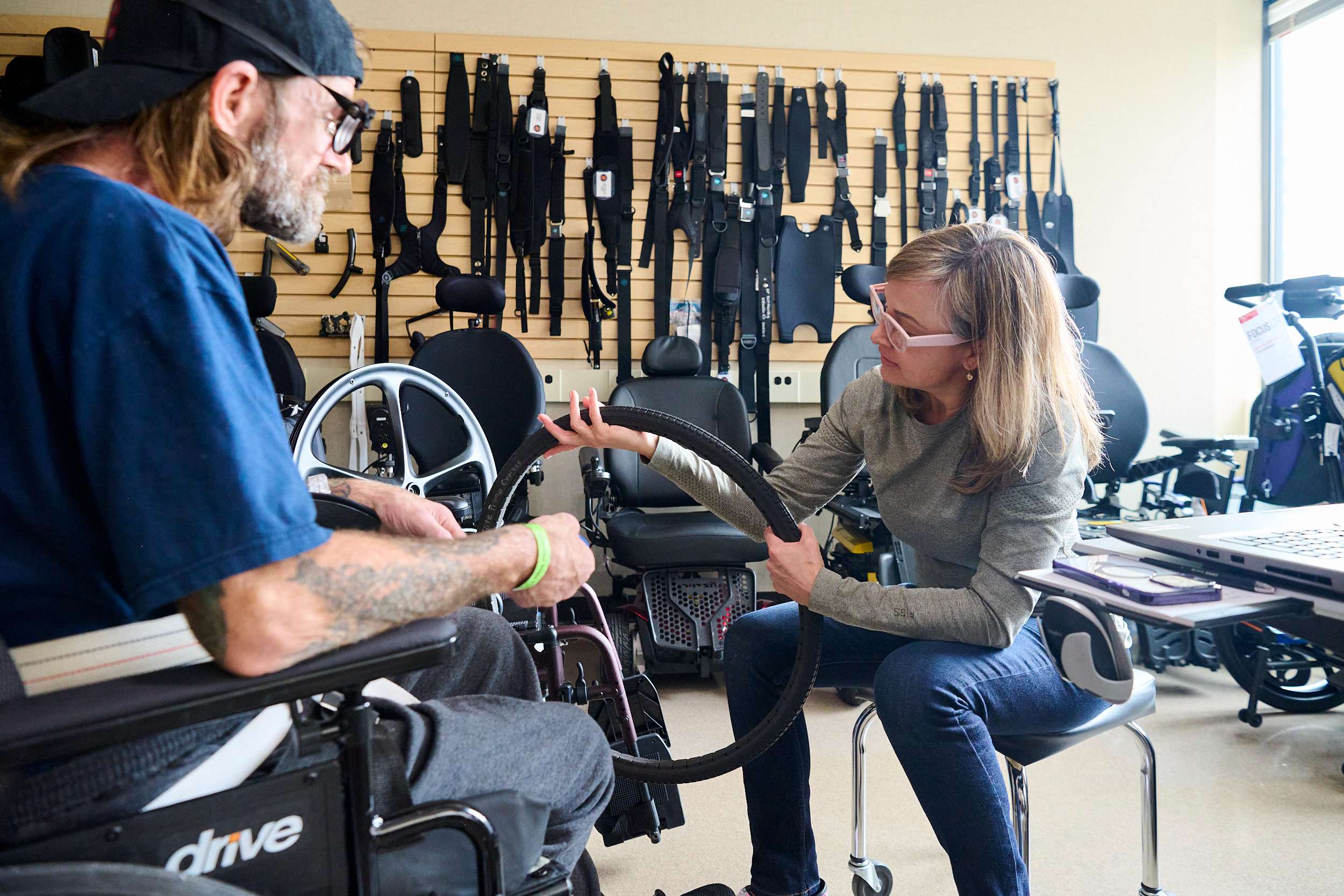 A woman assists a man in a wheelchair by holding a wheelchair tire. They are in a room filled with various wheelchair parts and accessories on the wall behind them.