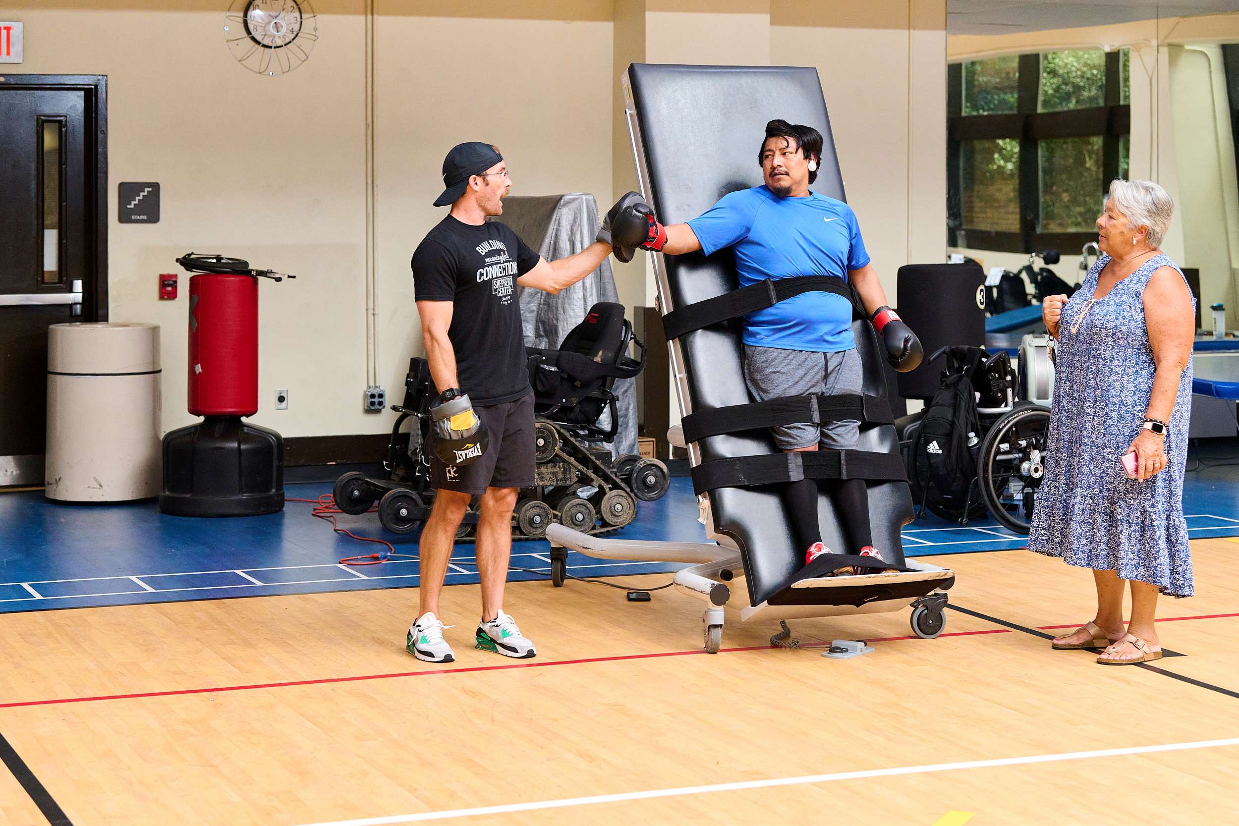 A man in sportswear assists another man who is strapped to a tilted workout bench, as a woman looks on. The setting is a gym with exercise equipment visible in the background.