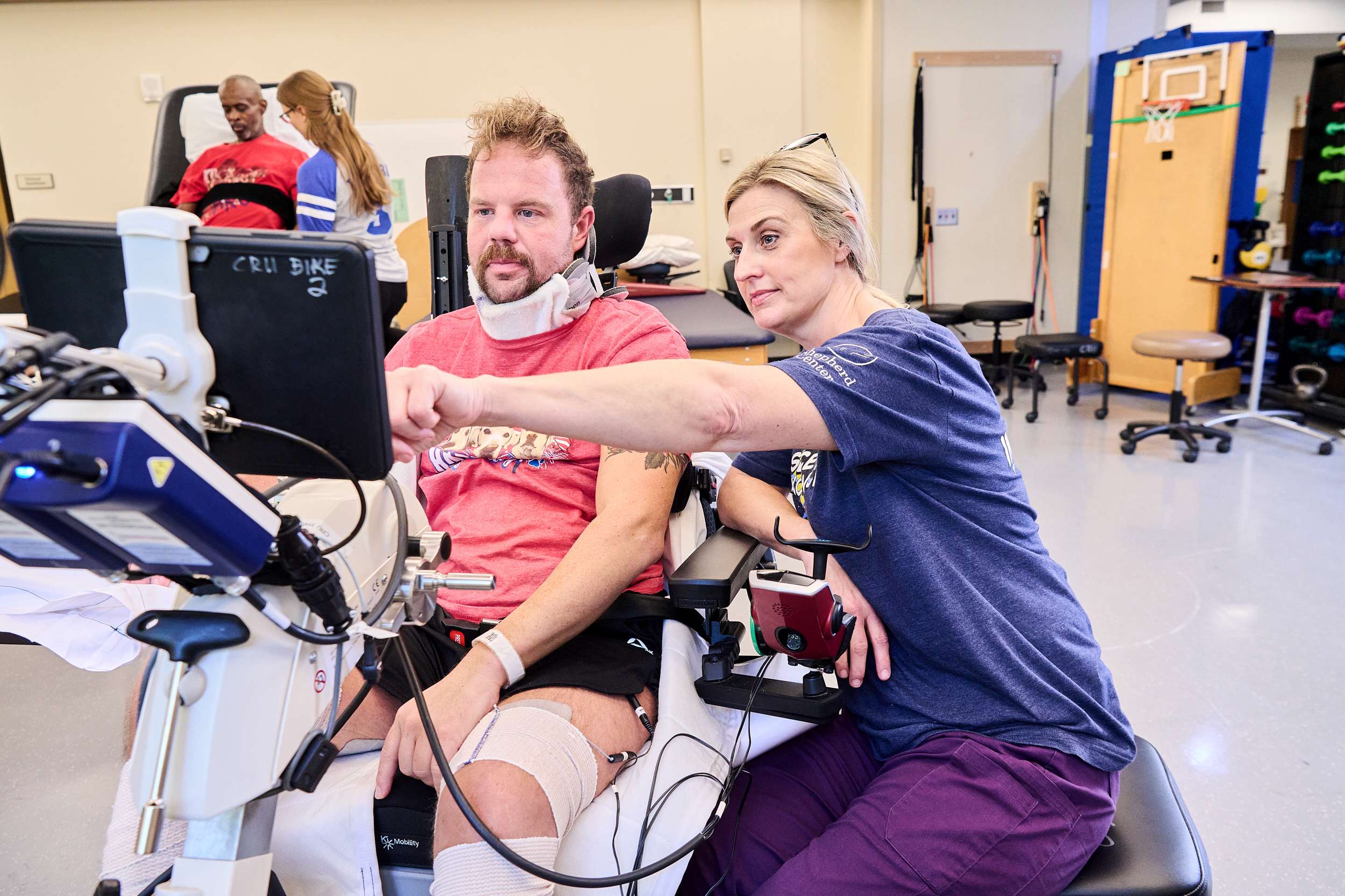 A man undergoing physical therapy sits in a specialized wheelchair, focused on a screen. A therapist beside him points at the screen, guiding him. Both are in a rehabilitation center with various equipment in the background.