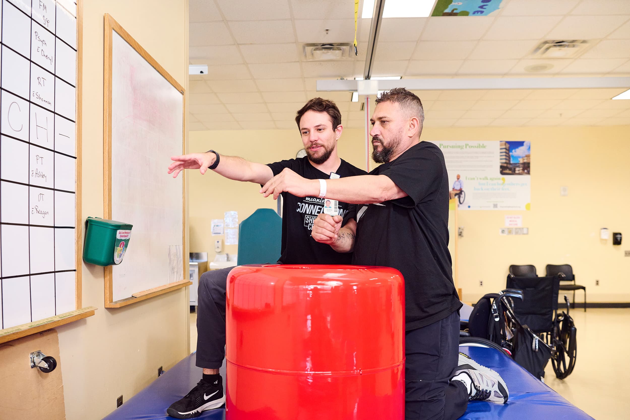 A physical therapist helps a man on a rehabilitation mat in a clinic. The therapist is guiding the patient's arm while holding it, and they appear to be engaged in an exercise. A large red cylinder is in front of them, and there is a wheelchair nearby.