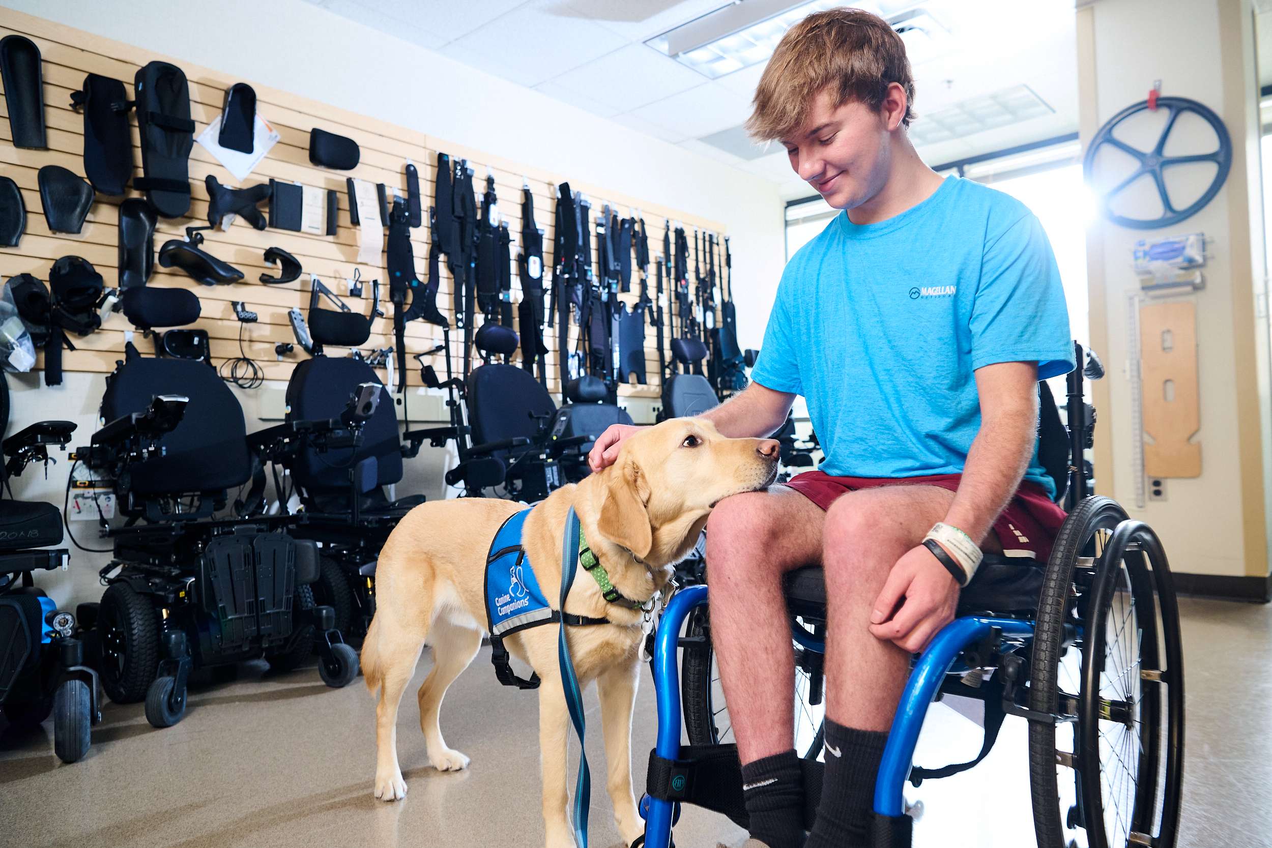A young person in a wheelchair pets a service dog in a room filled with mobility equipment. The person wears a light blue shirt and looks at the dog affectionately, while the dog wears a blue vest.