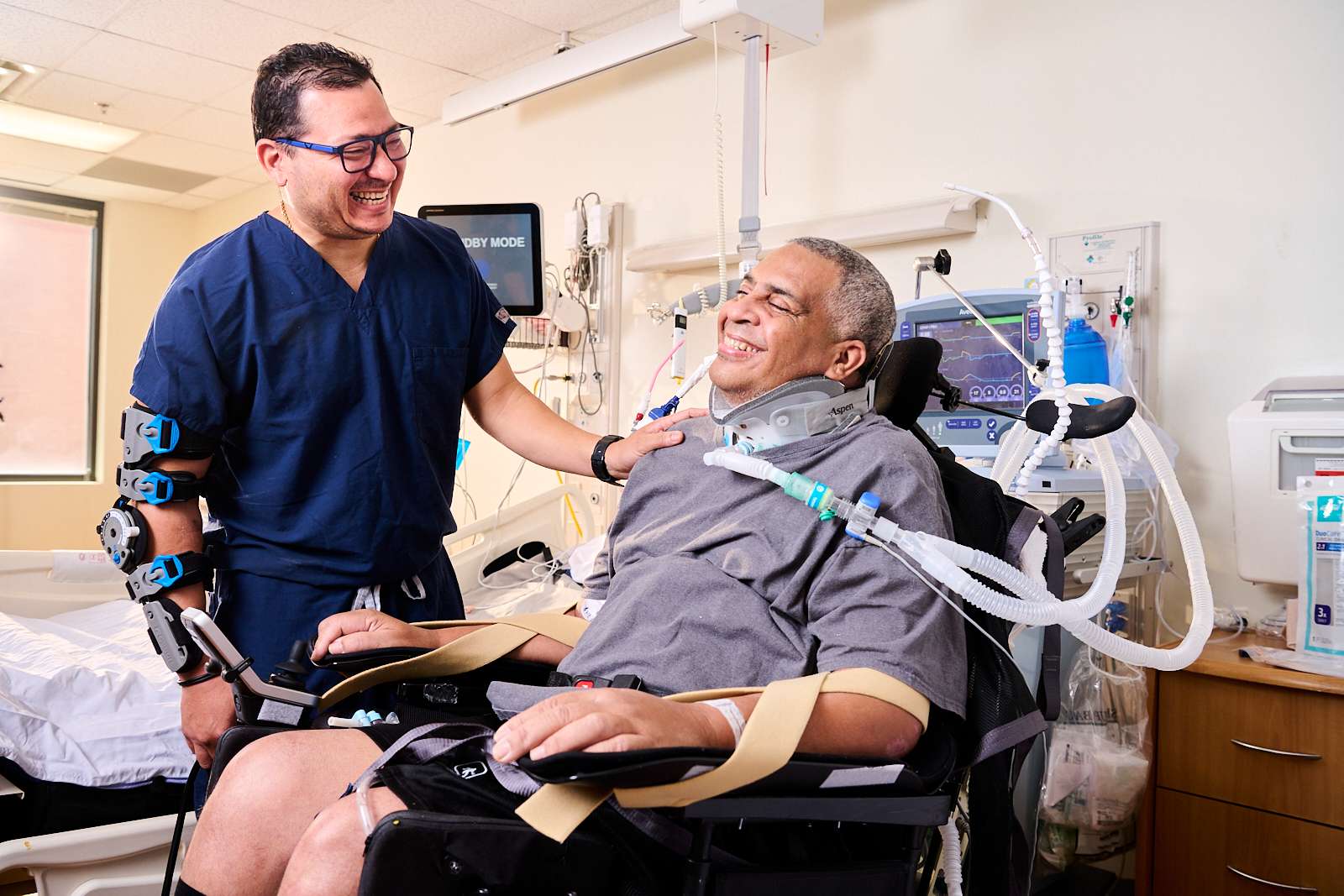 A healthcare professional in blue scrubs smiles and talks with a patient sitting in a wheelchair. The patient is wearing medical devices and appears to be in a hospital room. Medical equipment is visible in the background.