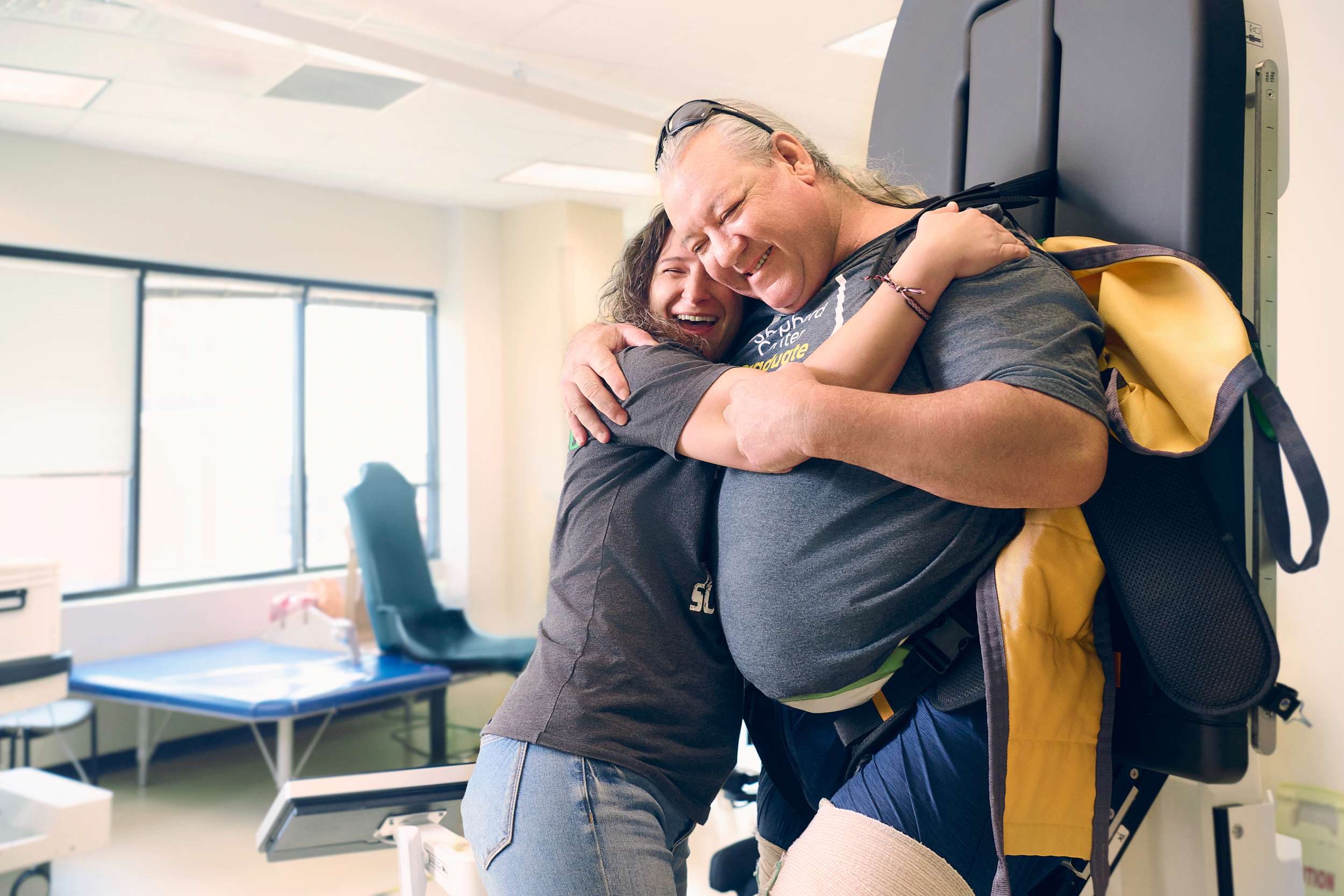 A person in a gray t-shirt hugs another person who is smiling and leaning against a medical support device in a bright room. The setting appears to be a clinic or rehabilitation center.