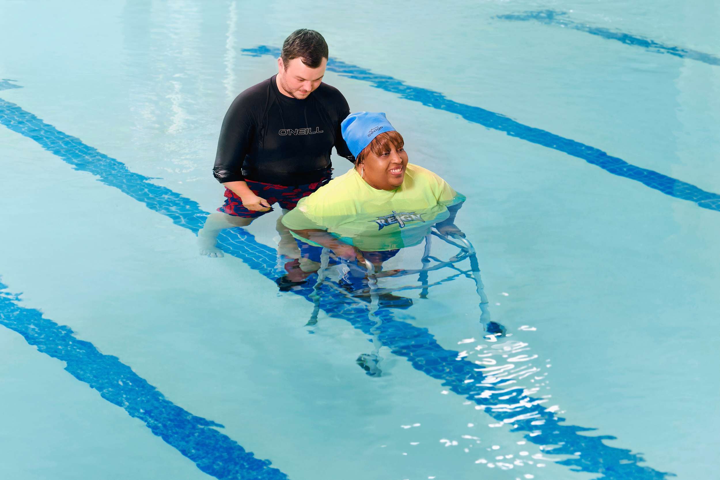 A person assists another who is using a walker for support in a swimming pool. The water is clear, and blue lines are visible on the pool floor. Both individuals are partially submerged and appear focused on the activity.