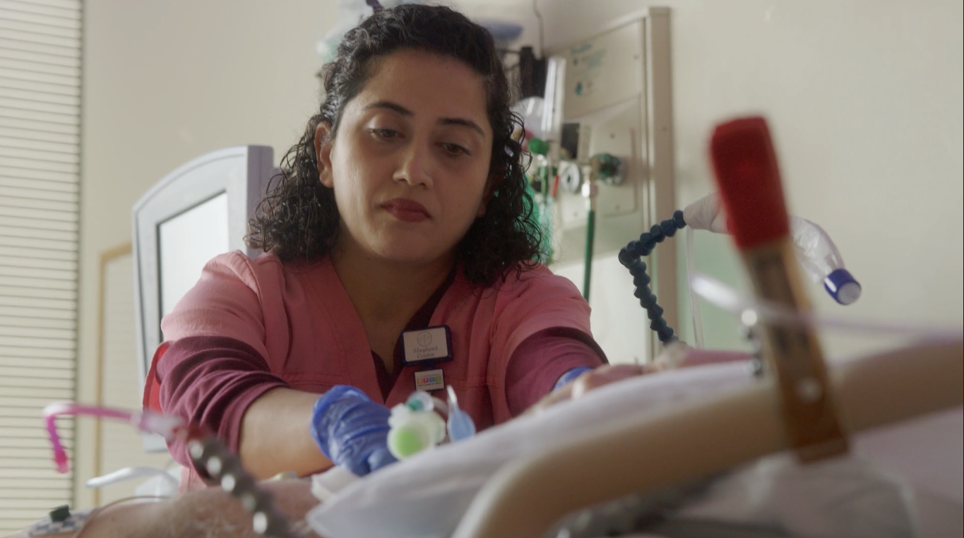 A healthcare professional in pink scrubs and blue gloves tends to a patient in a medical setting. She's focused on her task, with medical equipment visible in the foreground and background.