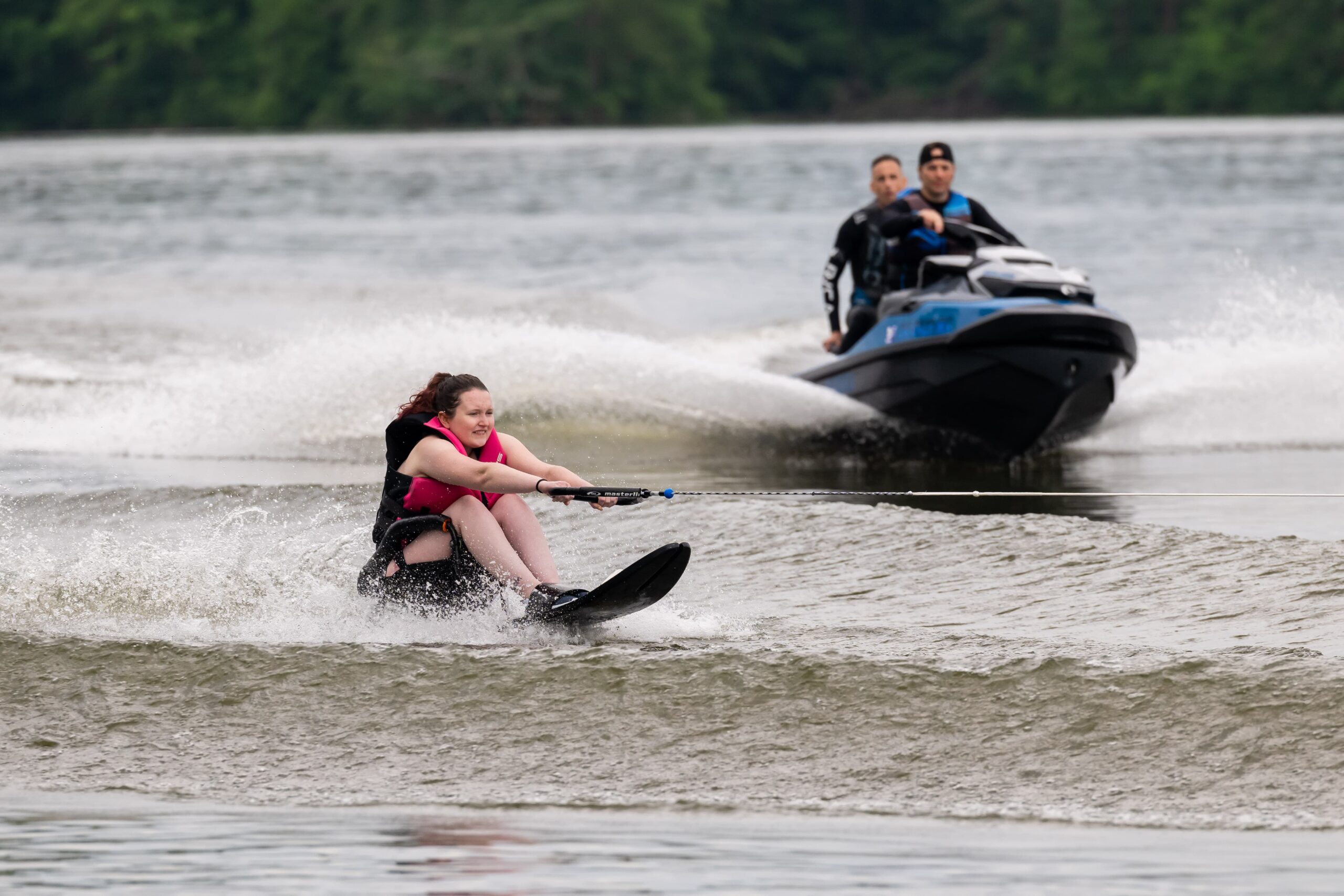 Person waterskiing on a lake, being pulled by a rope attached to a jet ski. Two people are riding the jet ski, steering it with water splashing around. The background is a wooded shoreline under an overcast sky.