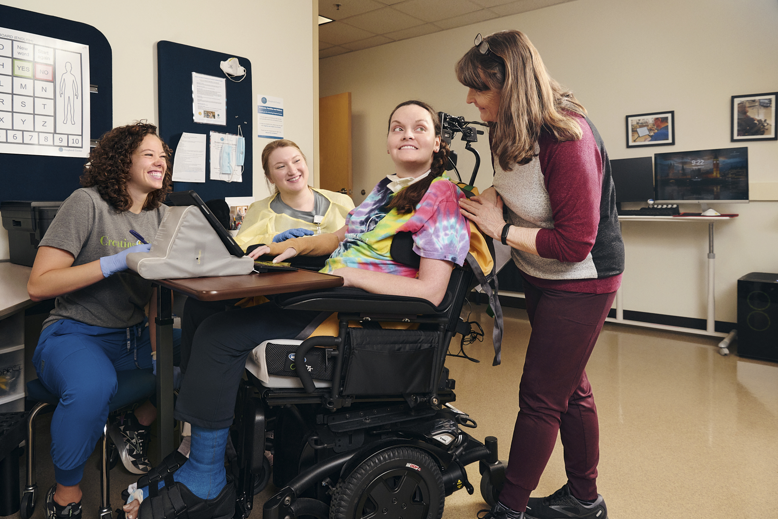 A person in a wheelchair smiles while two people on the left and one person on the right engage with them in a supportive setting. The room has a computer station and medical posters.