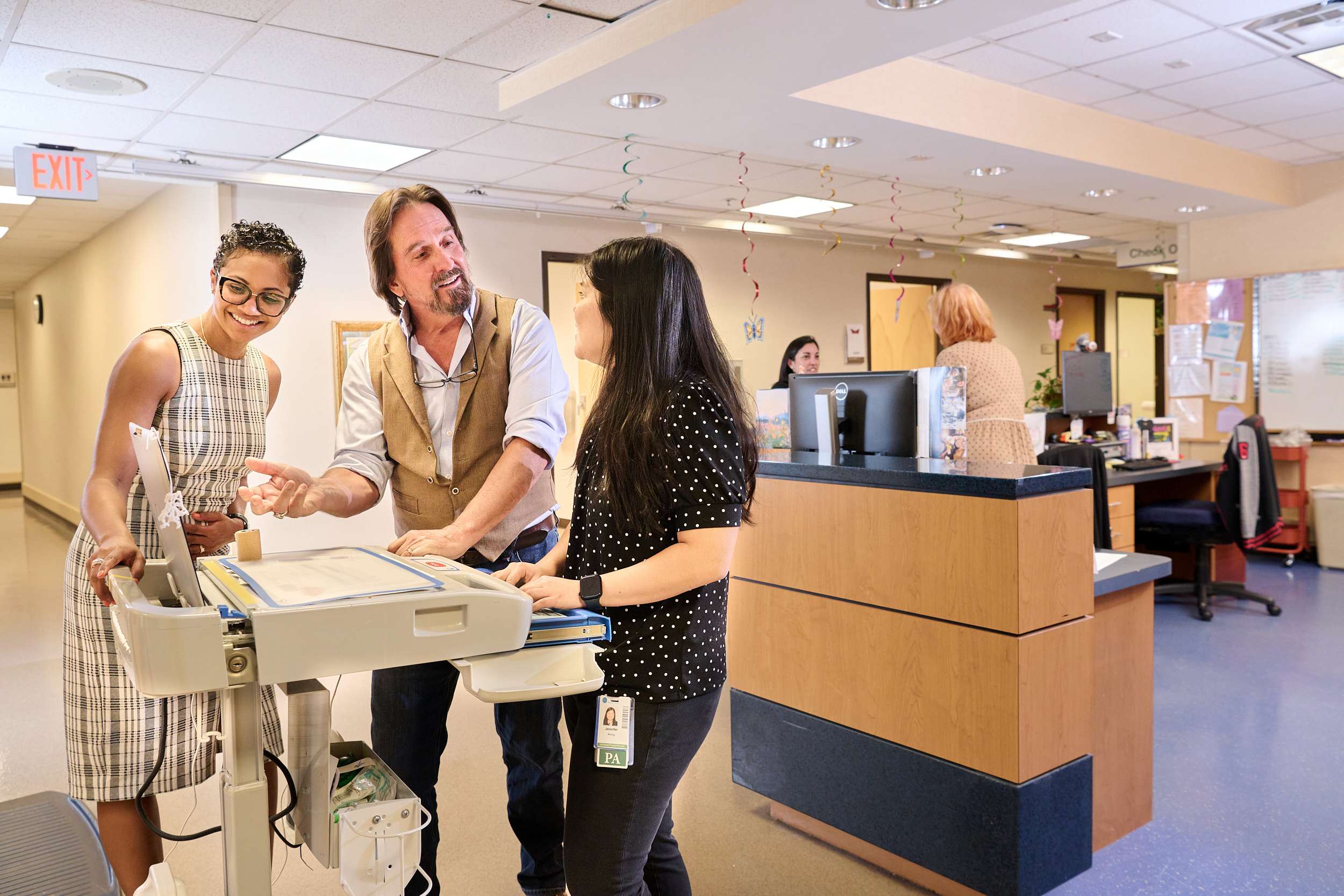 Three people, including two women and a man, stand smiling and conversing around a medical cart in a hospital reception area. A reception desk with a computer and another person are visible in the background.