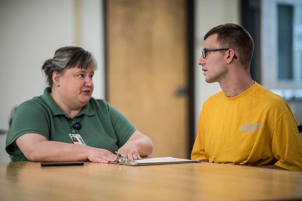 A woman and a man are sitting at a table in an office setting. The woman, wearing a green shirt, is talking to the man, who is wearing a yellow shirt and glasses. A clipboard is on the table in front of them.