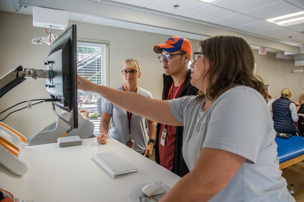 Three people stand around a computer screen in a workspace. One woman points at the monitor while explaining something to the other two. They are focused and engaged in the discussion.