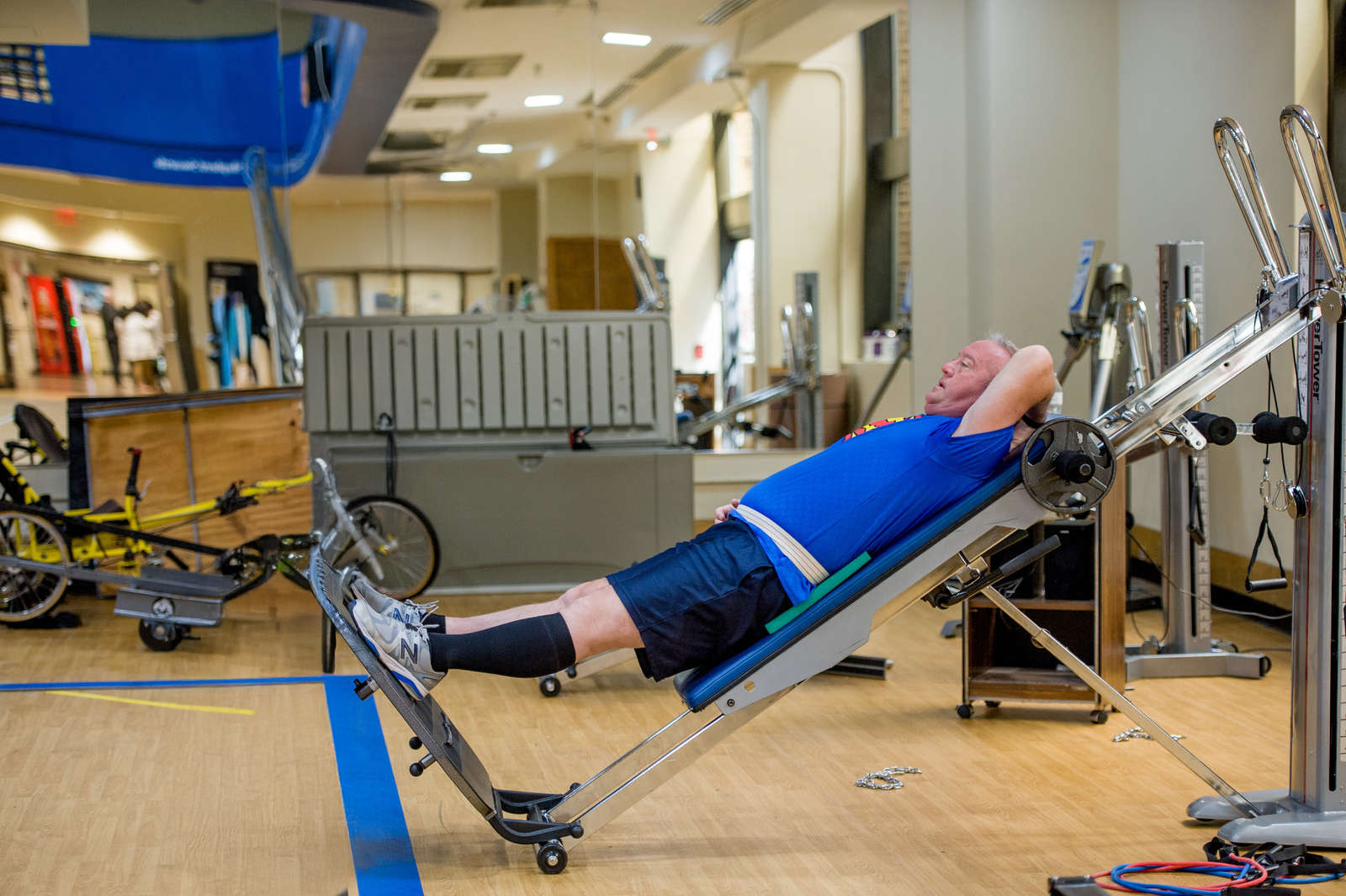 A man lies on an inclined exercise machine, wearing a blue shirt and knee-high socks, while a trainer in a black shirt and track pants stands beside him. The gym is equipped with various fitness equipment.