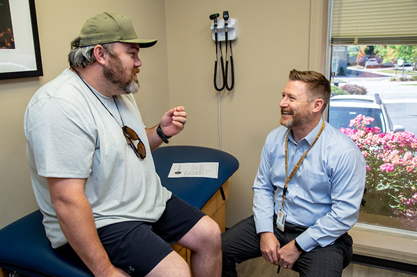 A bearded man in a cap and casual clothes sits on an examination table, animatedly speaking with a smiling man in a light blue shirt and lanyard. They are in a medical office with a window and flowers outside.