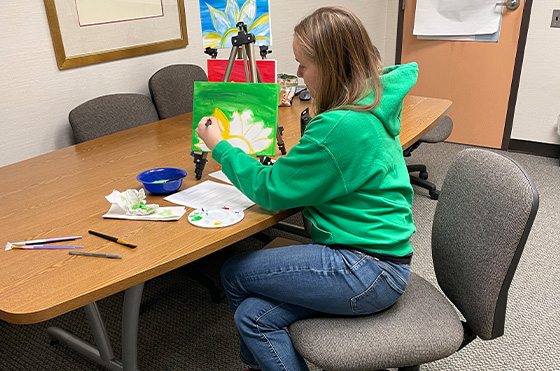 A person in a green hoodie sits at a wooden table, focusing on painting a flower on a small canvas. Various art supplies, including a palette and brushes, are scattered on the table. A doorway and another painting are visible in the background.