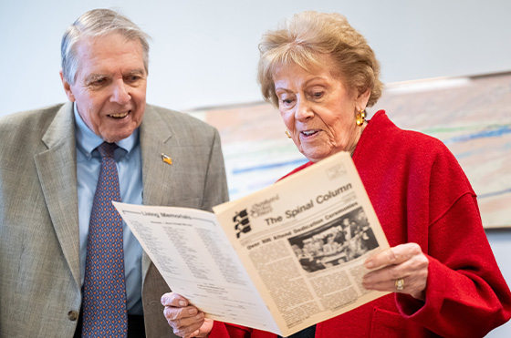 An elderly man and woman are standing together. The woman, wearing a red coat, is holding and reading a newspaper titled 