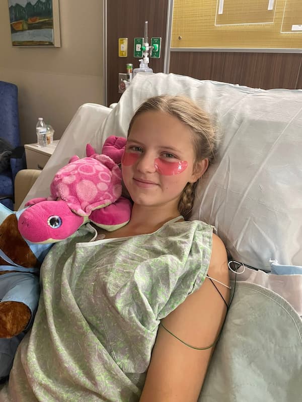 A girl with light hair in braids is lying in a hospital bed, wearing a hospital gown. She has pink patches under her eyes and is holding a pink stuffed turtle, smiling gently at the camera.