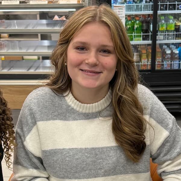 A young woman with wavy, light brown hair sits in a store, smiling while holding a cone of ice cream.