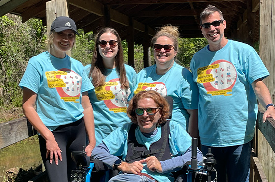 Five people wearing matching blue T-shirts with colorful graphics stand together outside on a sunny day. Four are standing, and one person is in a wheelchair in front. They are smiling and appear to be enjoying the event.