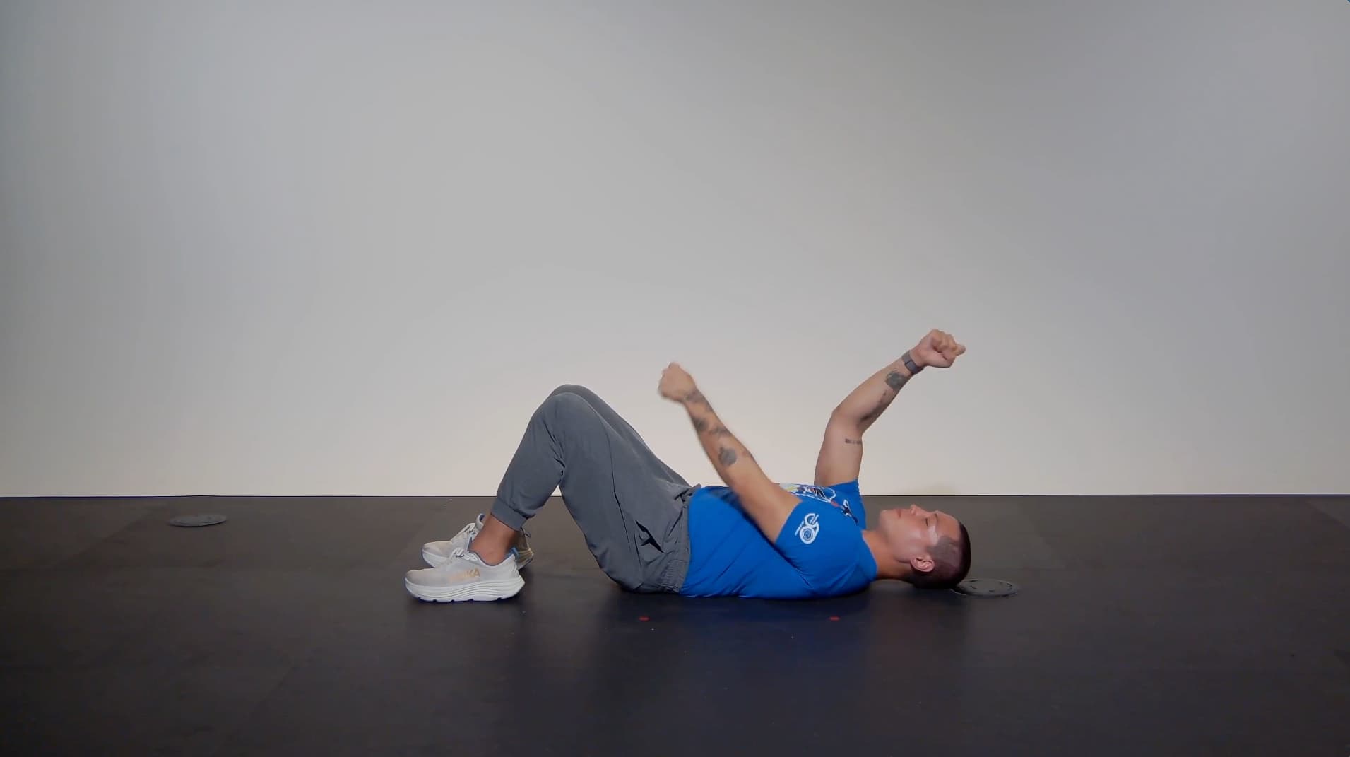 A person in a blue shirt, gray pants, and white sneakers lies on their back on a black floor with knees bent and arms raised, demonstrating an exercise movement in a minimal, well-lit space.