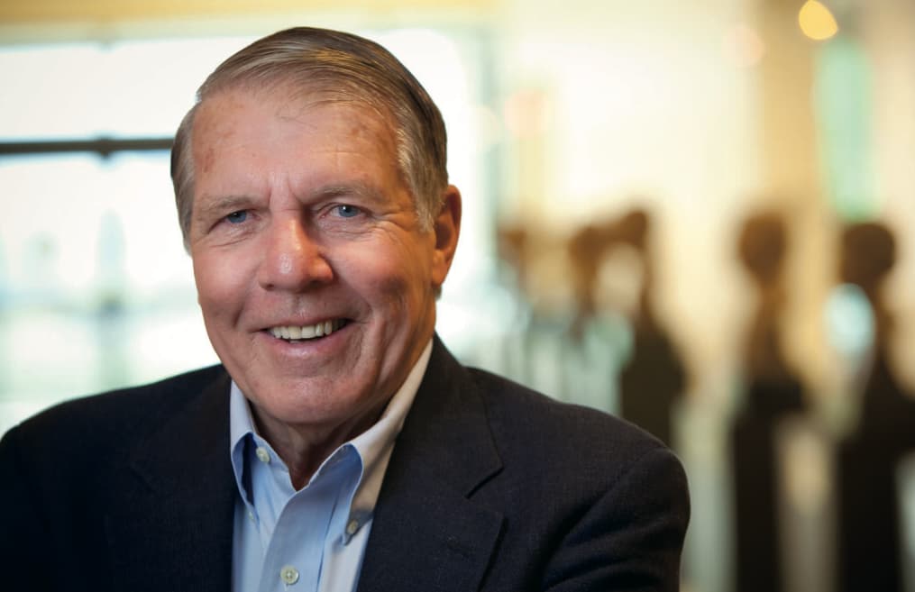Dr. Apple wears a dark suit jacket and light blue collared shirt, standing indoors with a blurred background of abstract shapes and warm lighting.