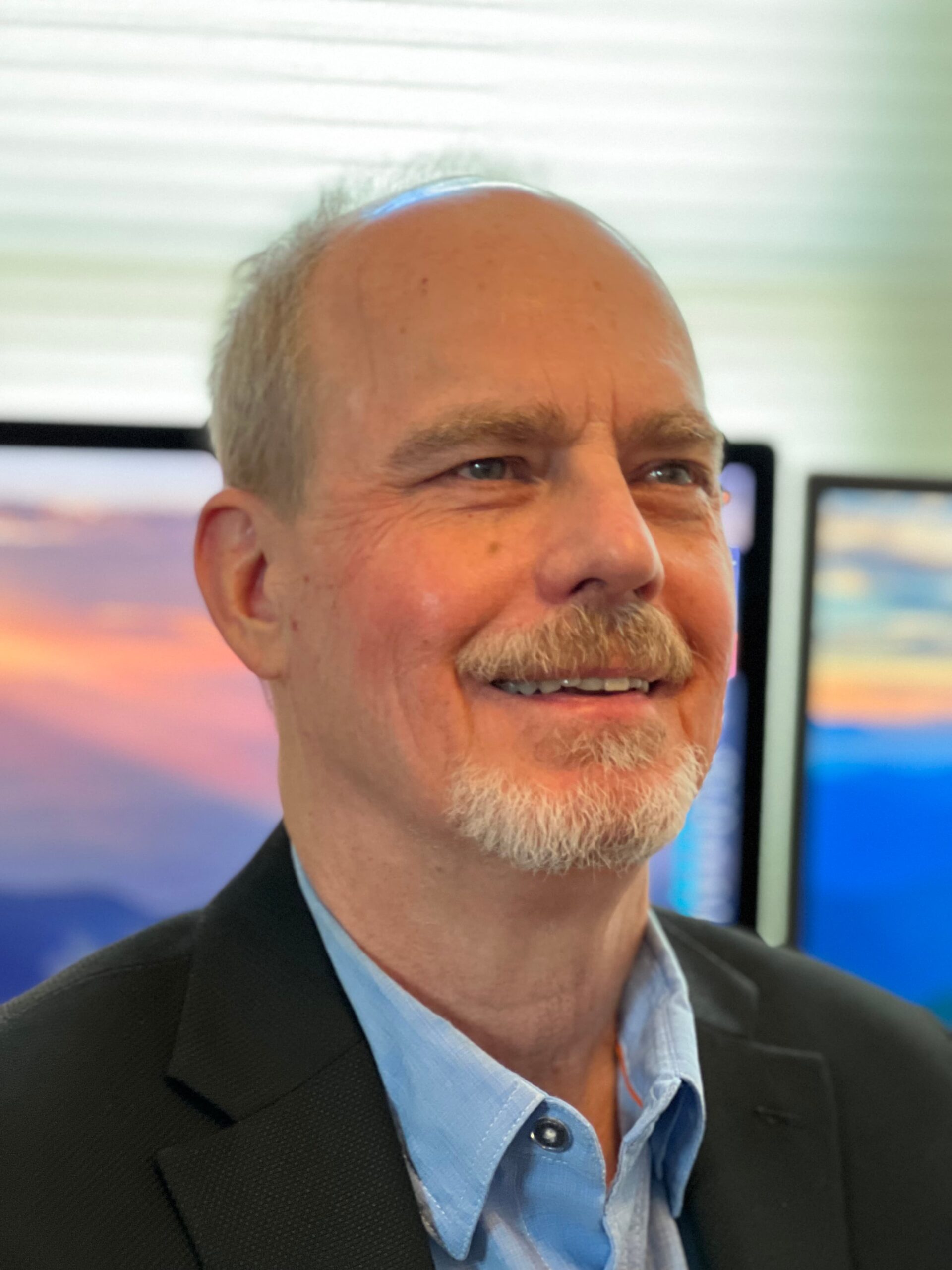 A middle-aged man with short gray hair and a goatee smiles slightly while wearing a navy zip-up jacket with red stripes. He stands outdoors with cloudy skies and distant mountains in the background.