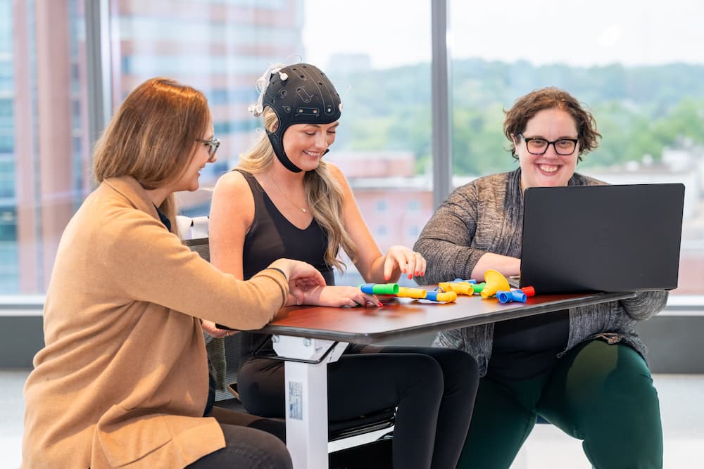 Three women sit at a table; one wears a brainwave cap and handles colorful plastic shapes, while another woman sits with a laptop, and the third observes and smiles. Large windows show buildings and greenery outside.