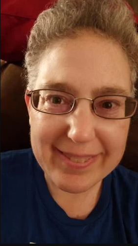 AURC member, Jennifer Beilis, a woman with short, curly gray hair wearing a dark blue shirt, seated against a dark background with a red cushion visible. The focus is on her upper torso and head area.