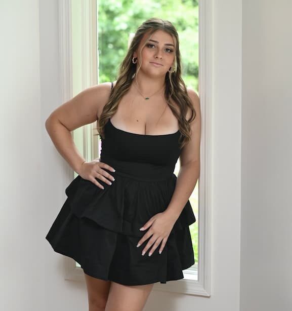 A woman with long brown hair, wearing a sleeveless black dress and hoop earrings, stands beside a bright window with greenery visible outside.