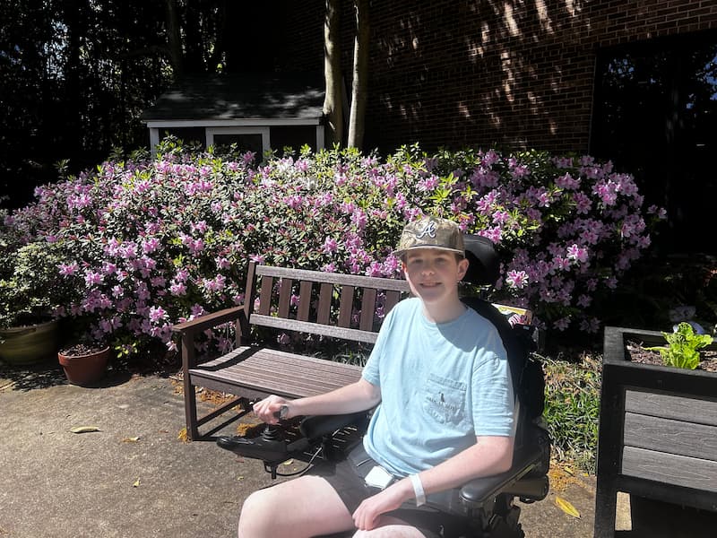 A teenage boy in a wheelchair smiles at the camera while sitting outside on a sunny day, with pink and purple azalea bushes and a wooden bench in the background.
