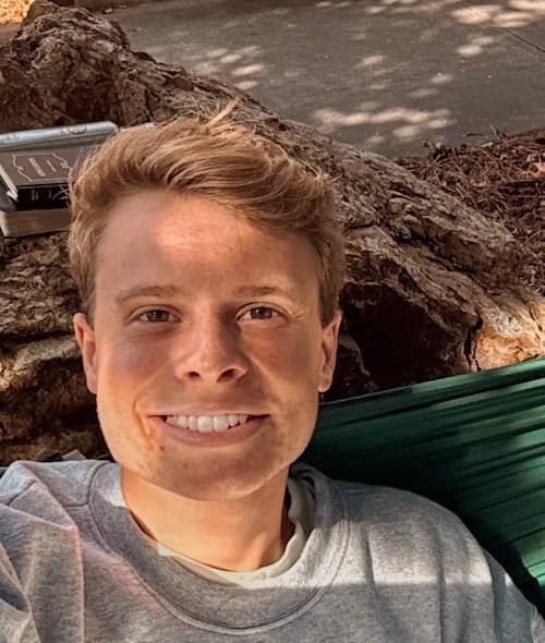A young person with short blonde hair smiles while reclining outdoors in a hammock, wearing a gray 