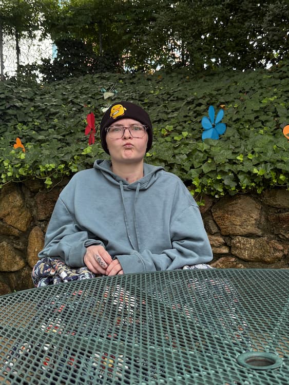 A person wearing glasses, a beanie, and a gray hoodie sits at a green metal table outdoors, in front of a stone wall covered with ivy and colorful flower decorations.