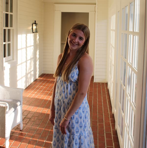 A young woman with long, light brown hair stands on a brick porch in a sleeveless, patterned blue and white dress, smiling at the camera.