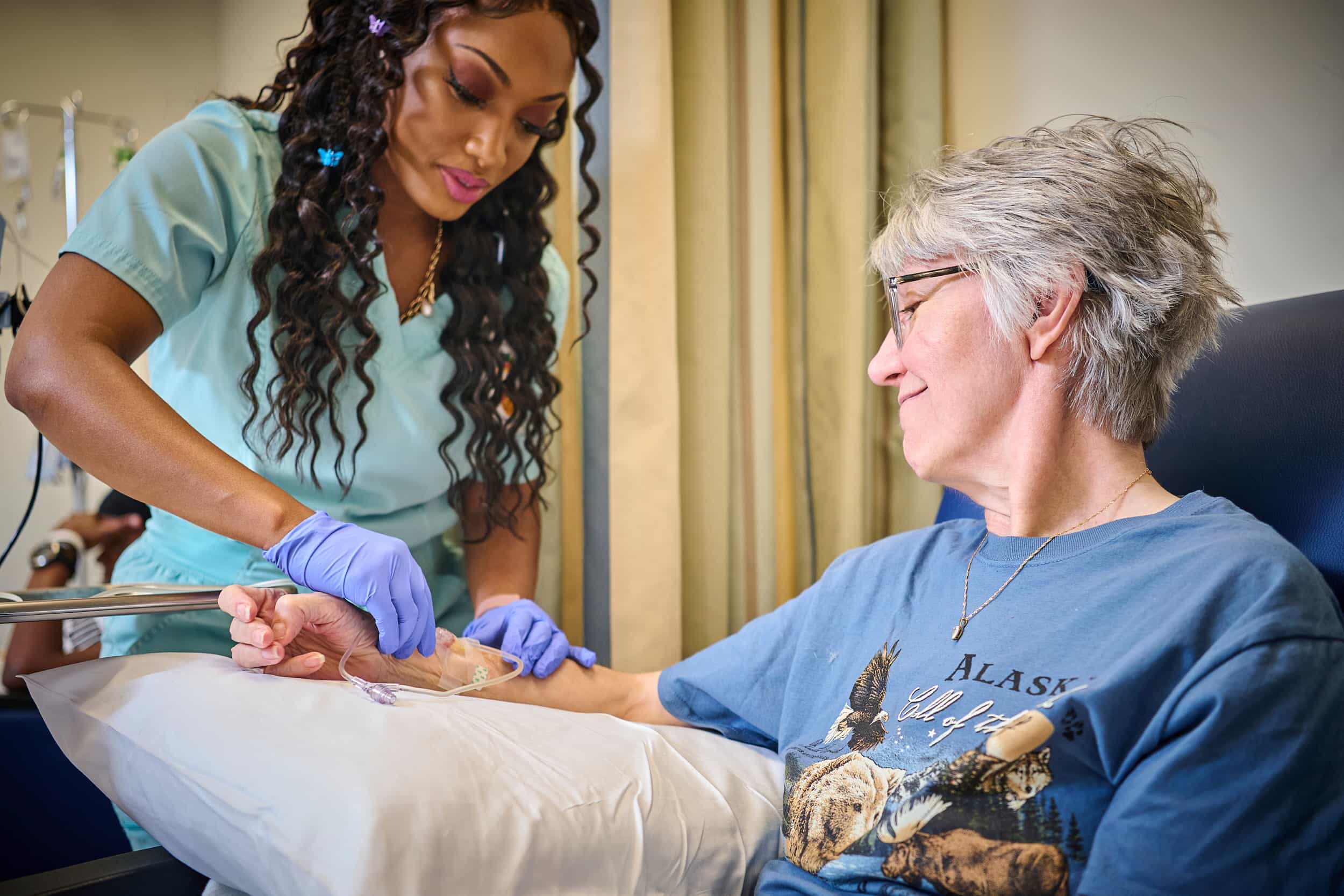 A nurse wearing gloves administers an IV to an older woman sitting in a chair, who is smiling and wearing glasses and a blue Alaska T-shirt. The woman rests her arm on a pillow.