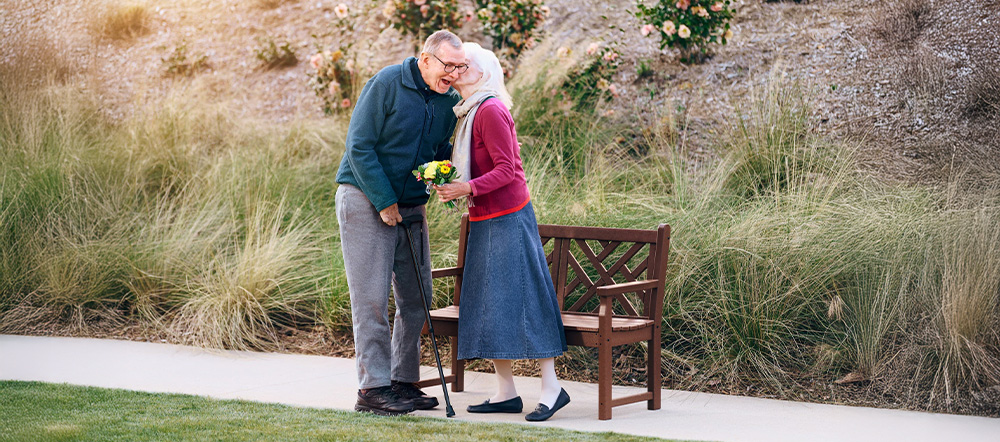 An older couple stands by a wooden bench in a garden. The woman, holding yellow flowers, leans in to kiss the man on the cheek. Both are smiling and dressed in warm clothes, with flowers blooming in the background.