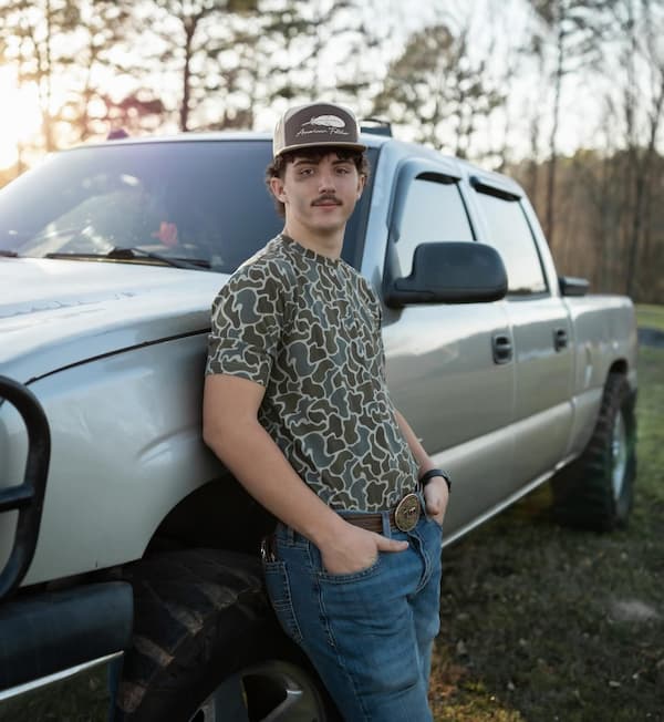 A young man in a patterned t-shirt, jeans, and a cap leans against the front of a large silver pickup truck, outdoors with trees and sunlight in the background.